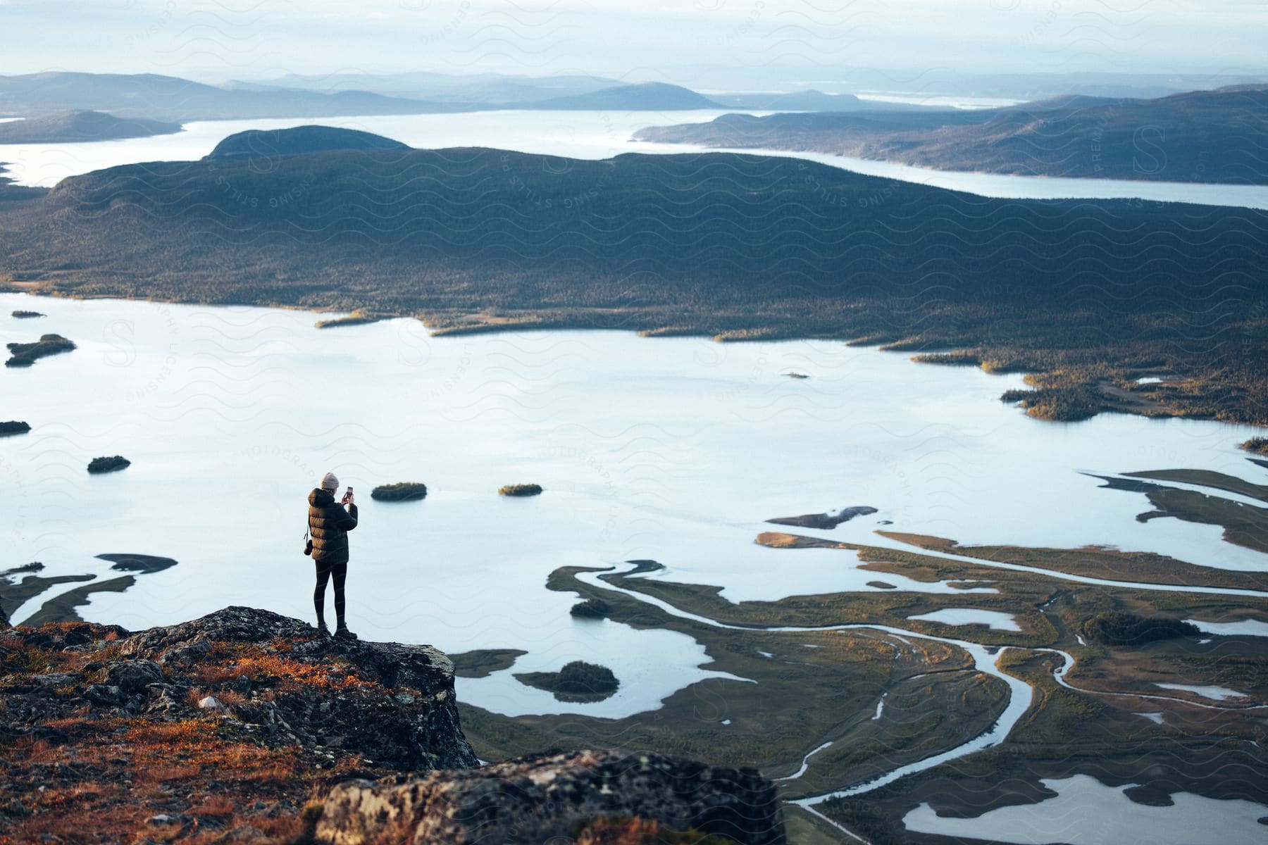 Person standing on a cliff overlooking a vast landscape with mountains and a lake.