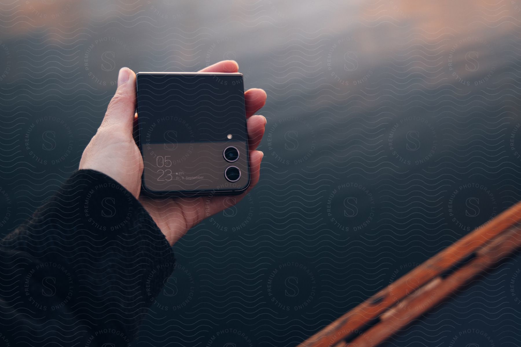 Stock photo of a person's hand holding a cell phone that is folded and showing the time in contrast with blue tones in the background