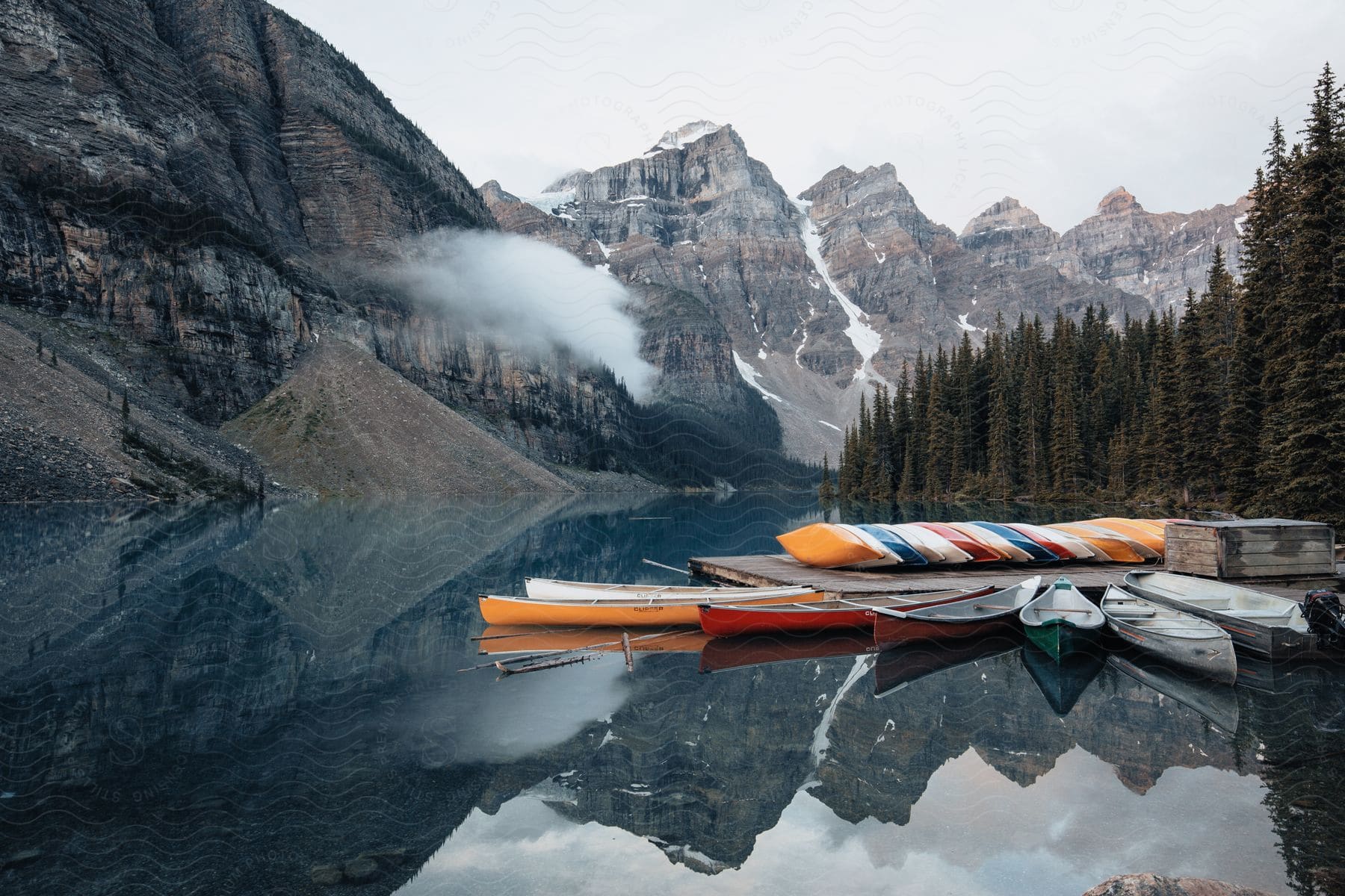 A group of colorful canoes sit next to a wooden dock on Moraine Lake on a cloudy day.