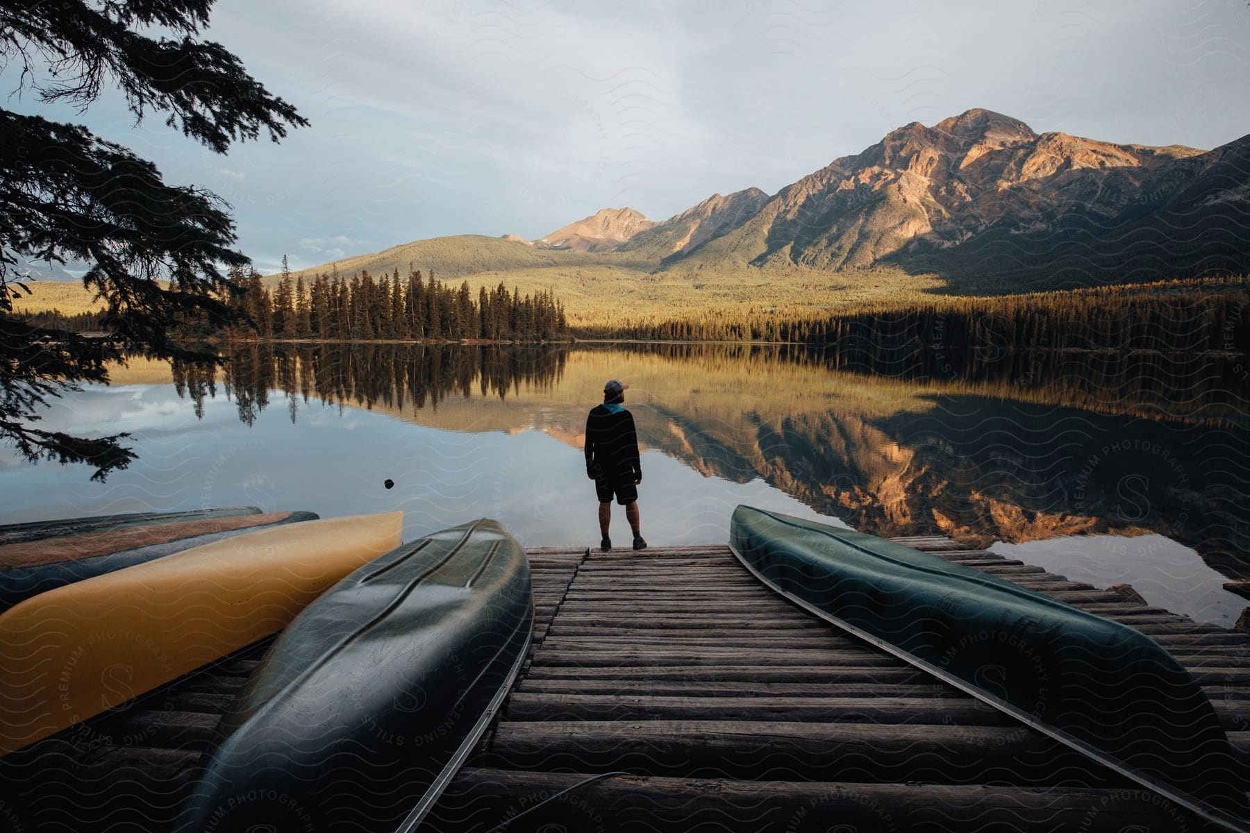 Man with his back standing at the end of a pier with canoes and looking at a calm lake with mountains and trees in the background.