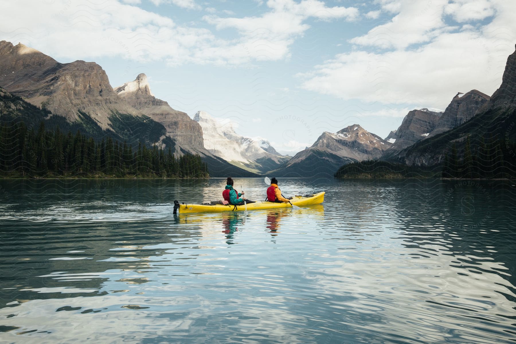 Two people are in a yellow kayak and paddling on a lake between mountains