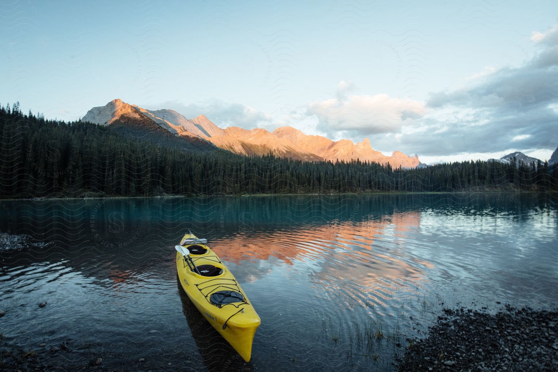 A yellow kayak on the shore of a tranquil lake, with majestic mountains and dense forests in the background, under a cloudy sky.