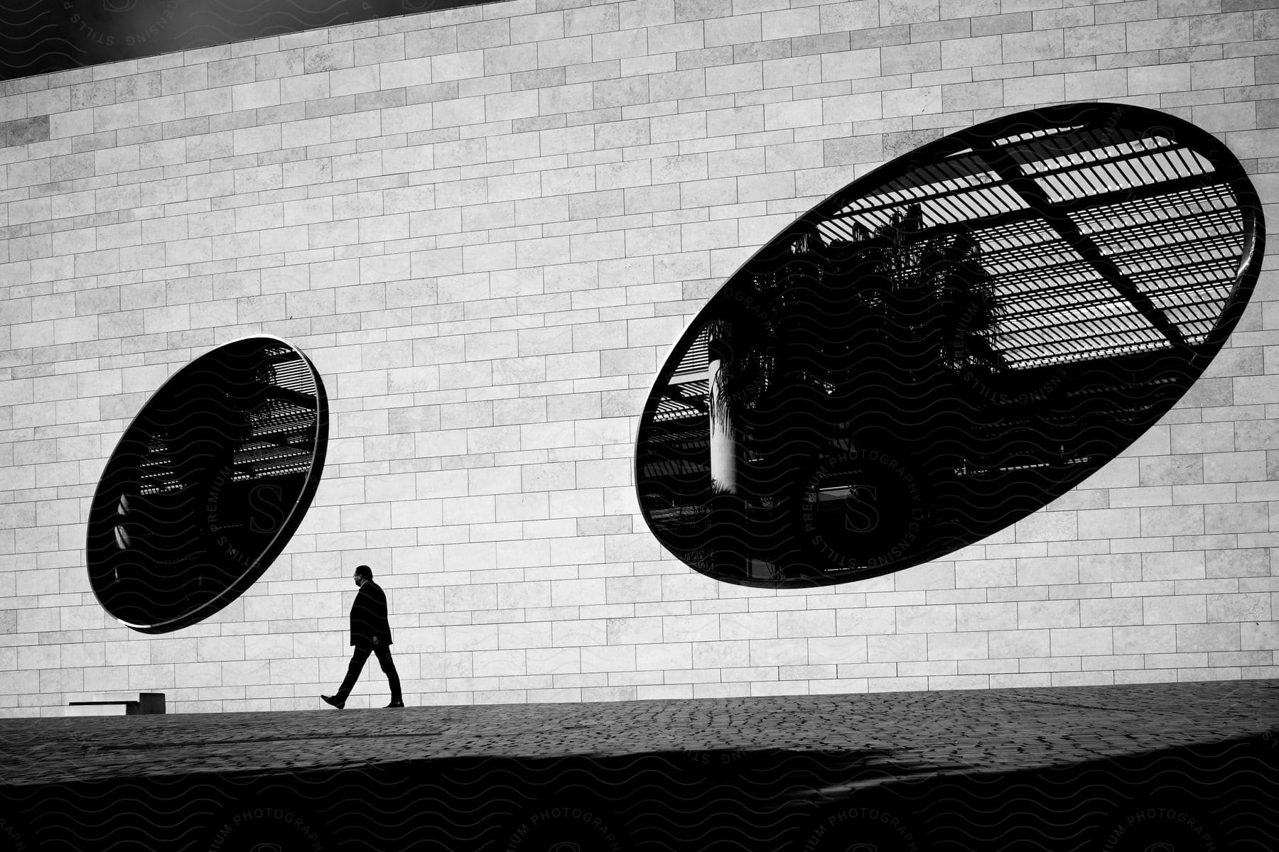A man walking near a white brick wall with two huge oval windows in black and white tones