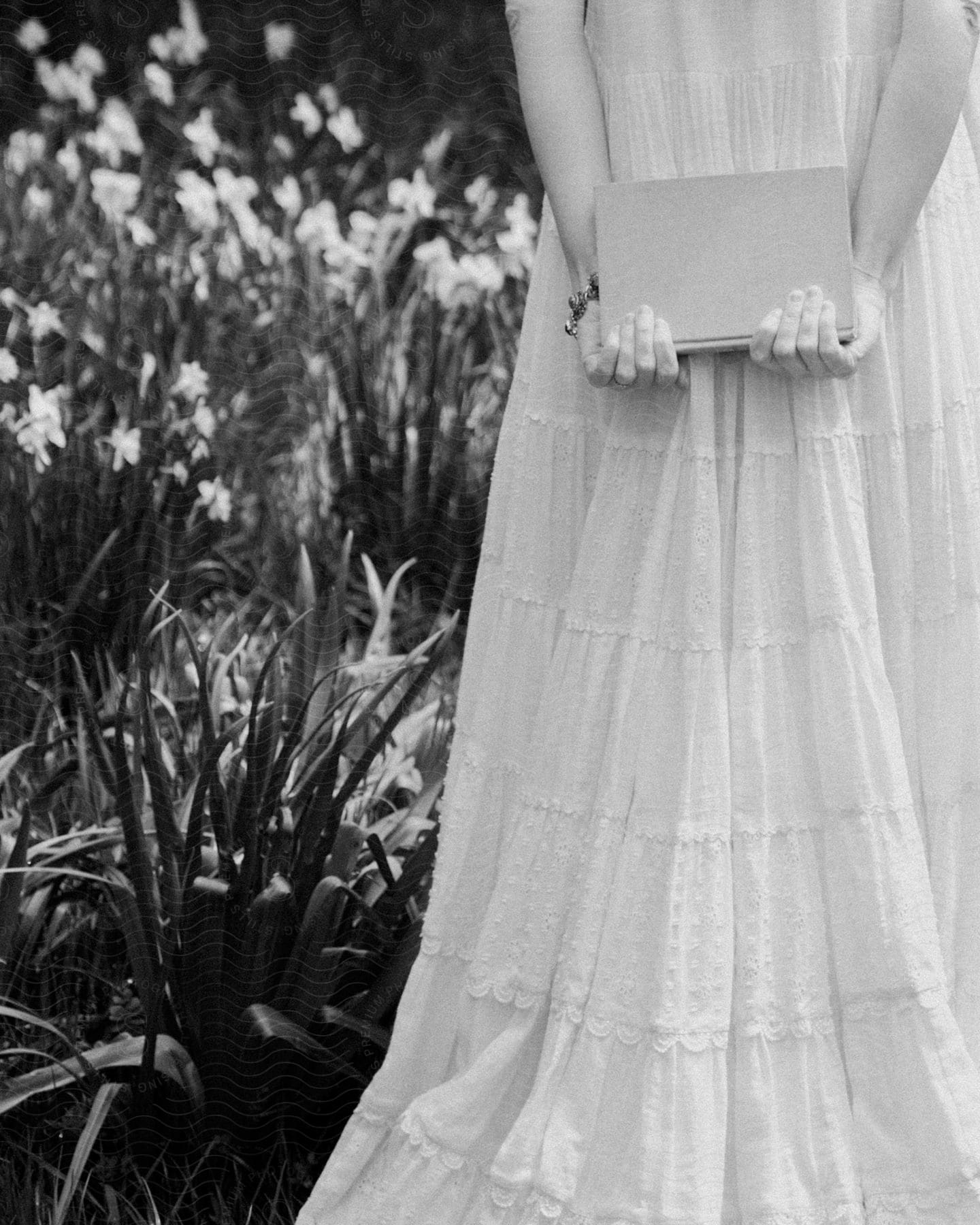 Woman with her back and arms behind her holding a book in a garden wearing a long white dress