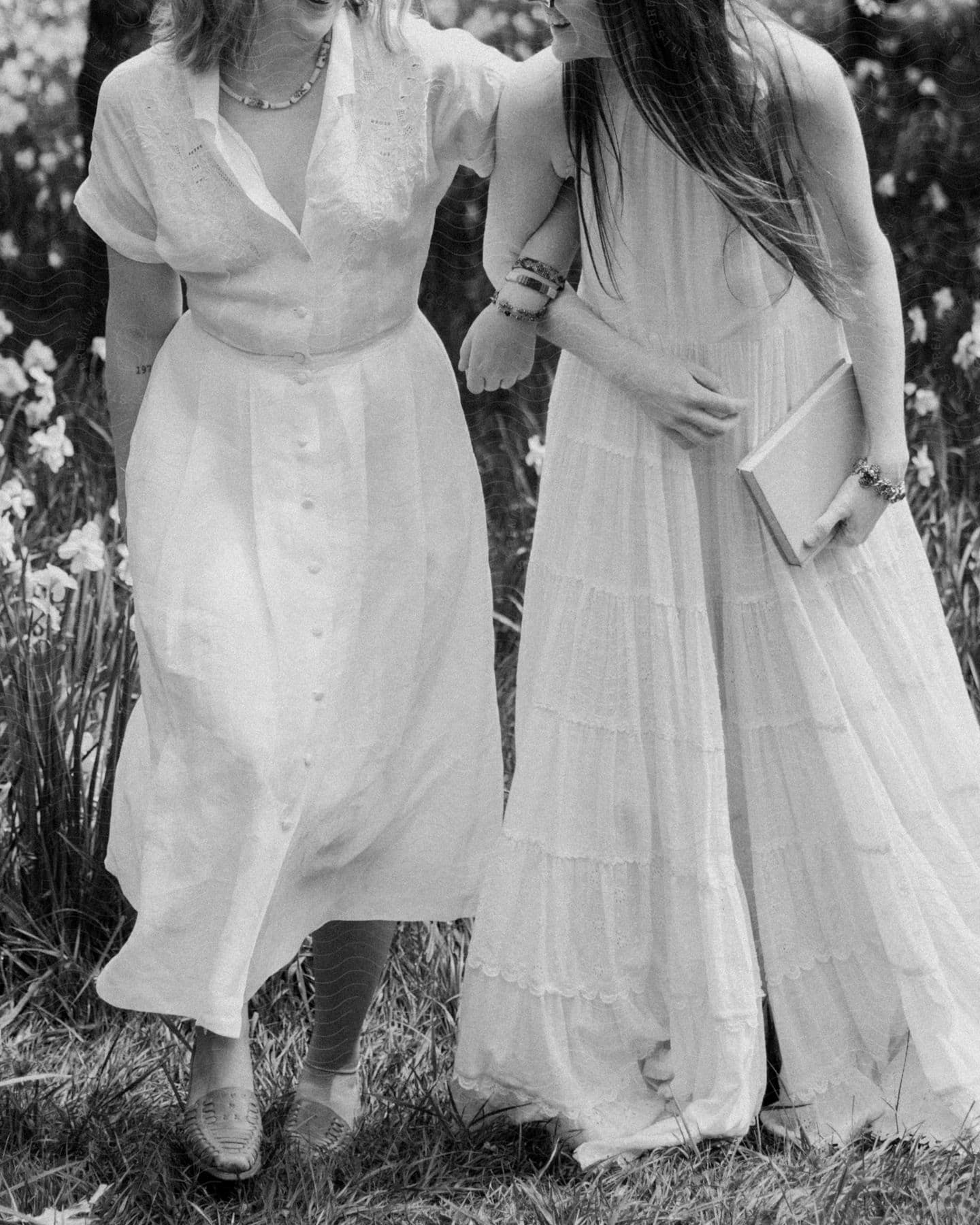 Two women arm in arm walking through the grass in a garden wearing long white dresses