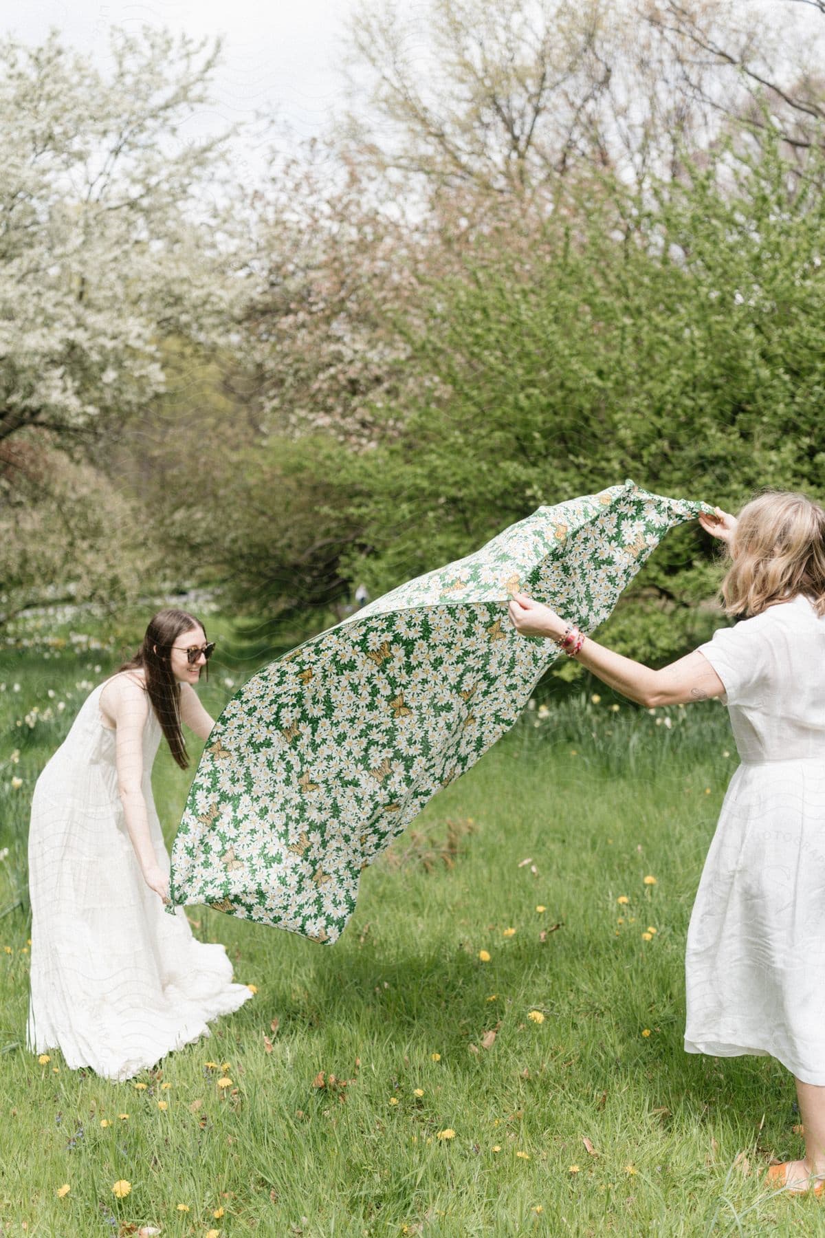 Two women in white dresses in an open-air garden spreading a Florida towel towards the grass