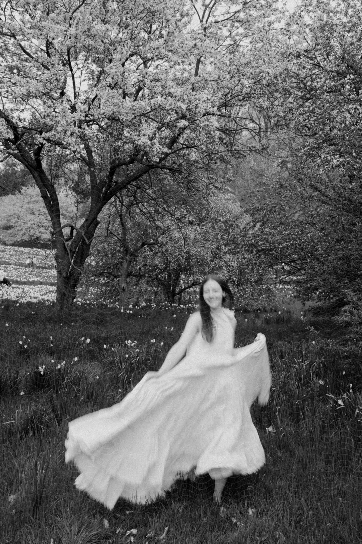 A woman in a long white dress is walking in a grass field and behind there is a tree with many flowers