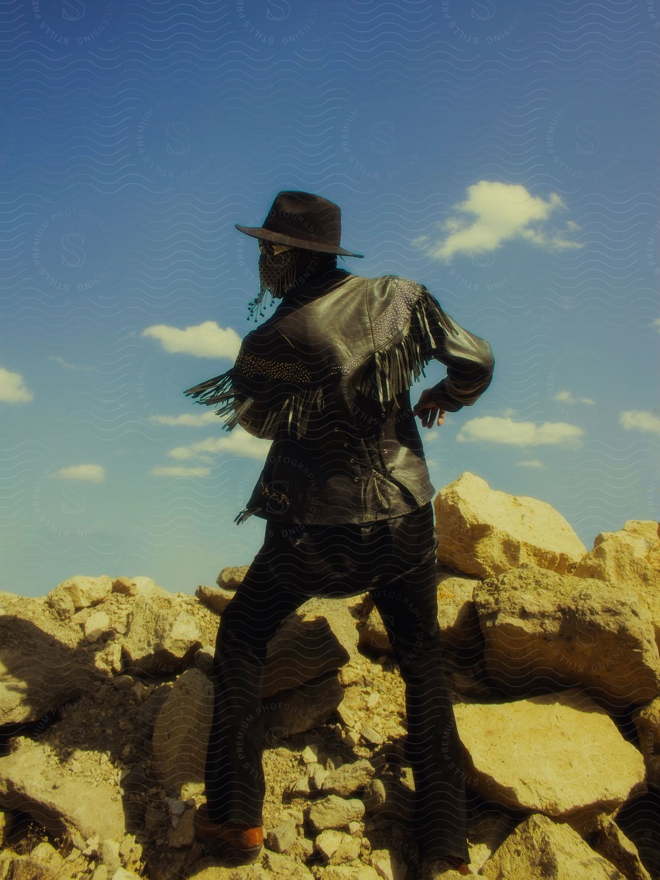 A person is on his back standing on rocks and wearing a leather jacket and a black hat and his jacket has small ribbons hanging from it