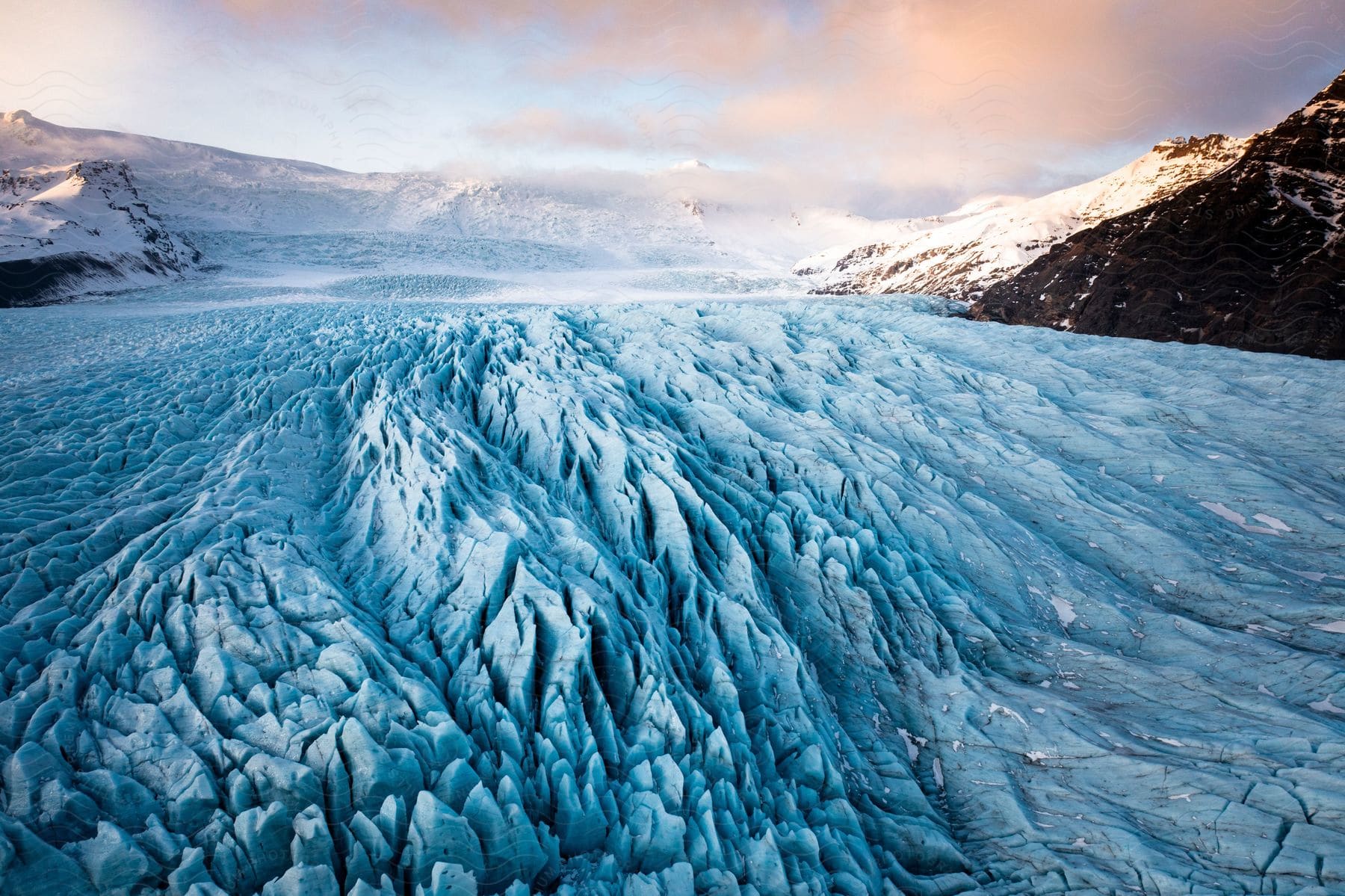 A breathtaking view of a vast, rugged glacier surrounded by snow-capped mountains under a partly cloudy sky.