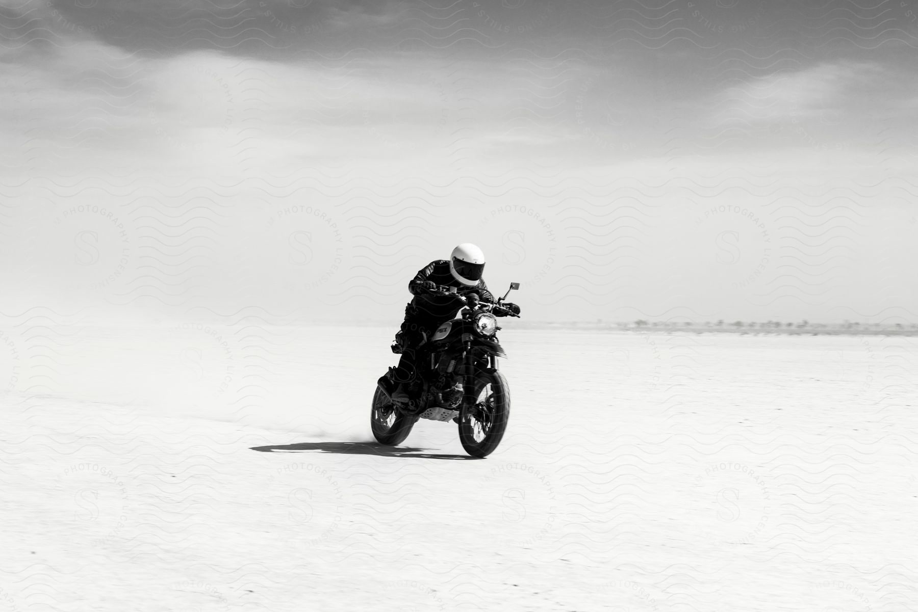 a biker wearing a white helmet, black jacket, and pants speeding up with a motorcycle in the desert.