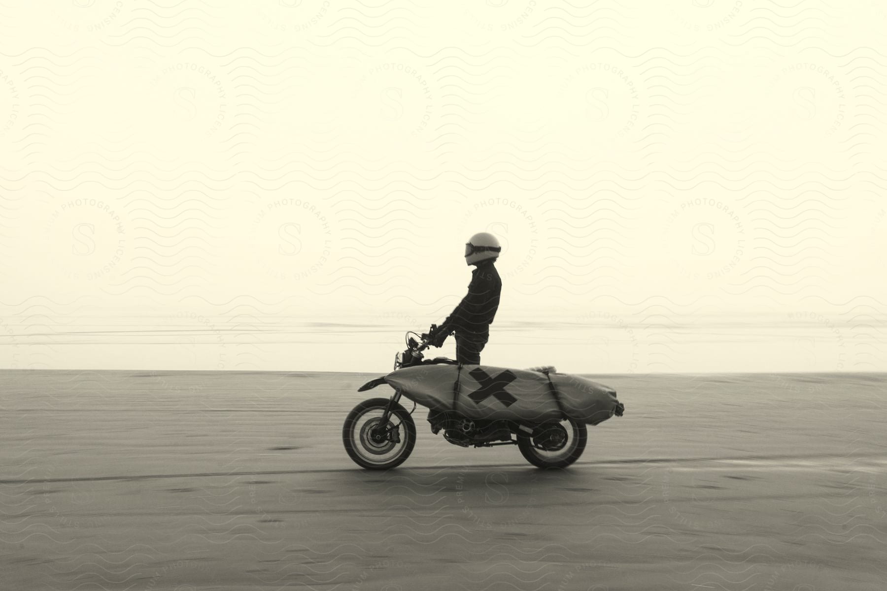 A person riding a motorcycle with a big X on the side, in the middle of a serene, open landscape.