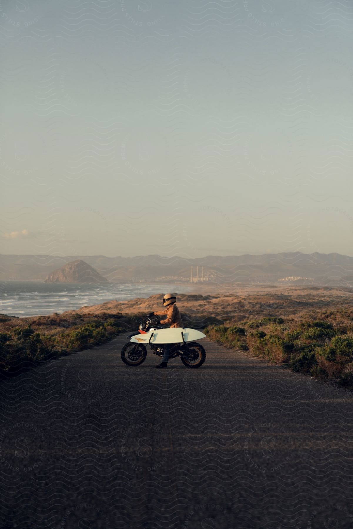 A biker sitting on a motorcycle holds a surfboard with the sea in the background.