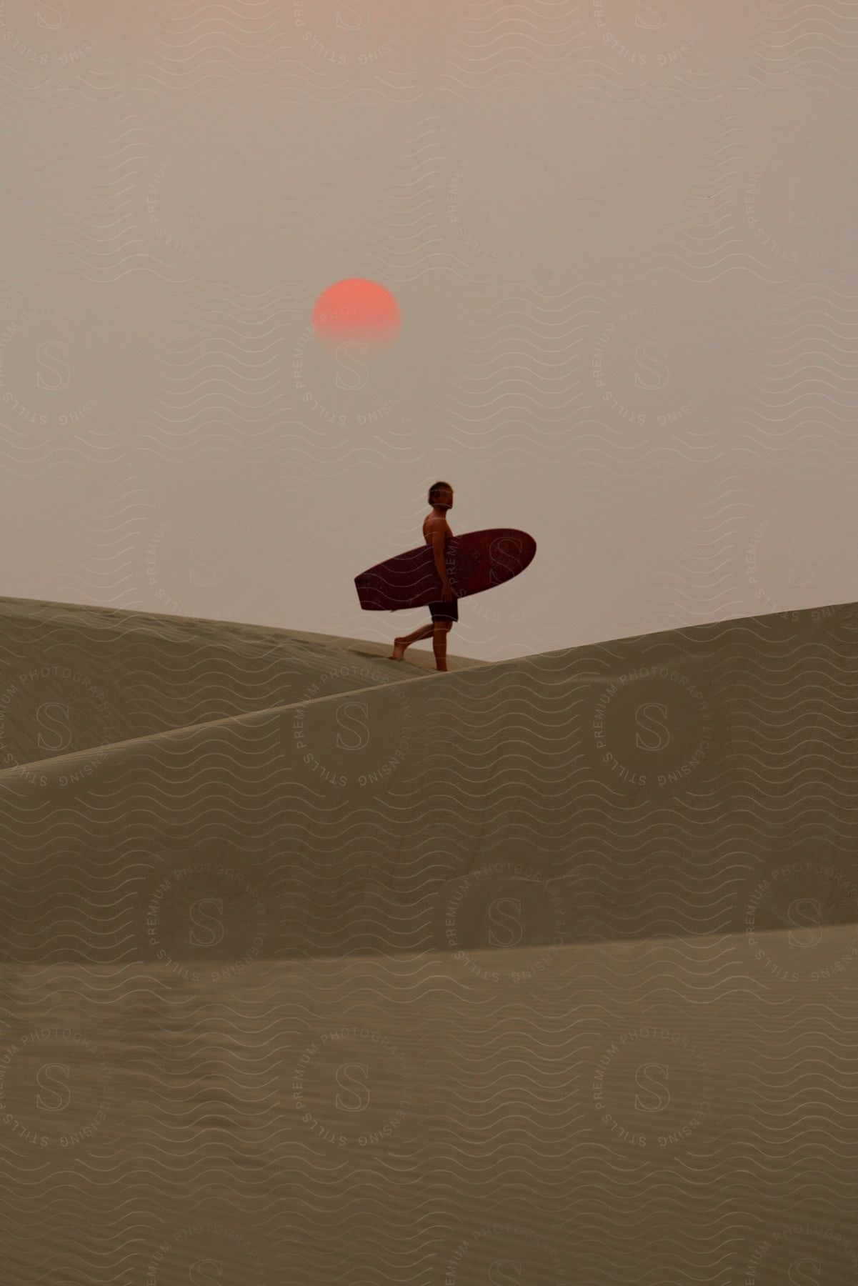 Stock photo of man wearing a shorts carries a surfboard in the desert with red moon in the background