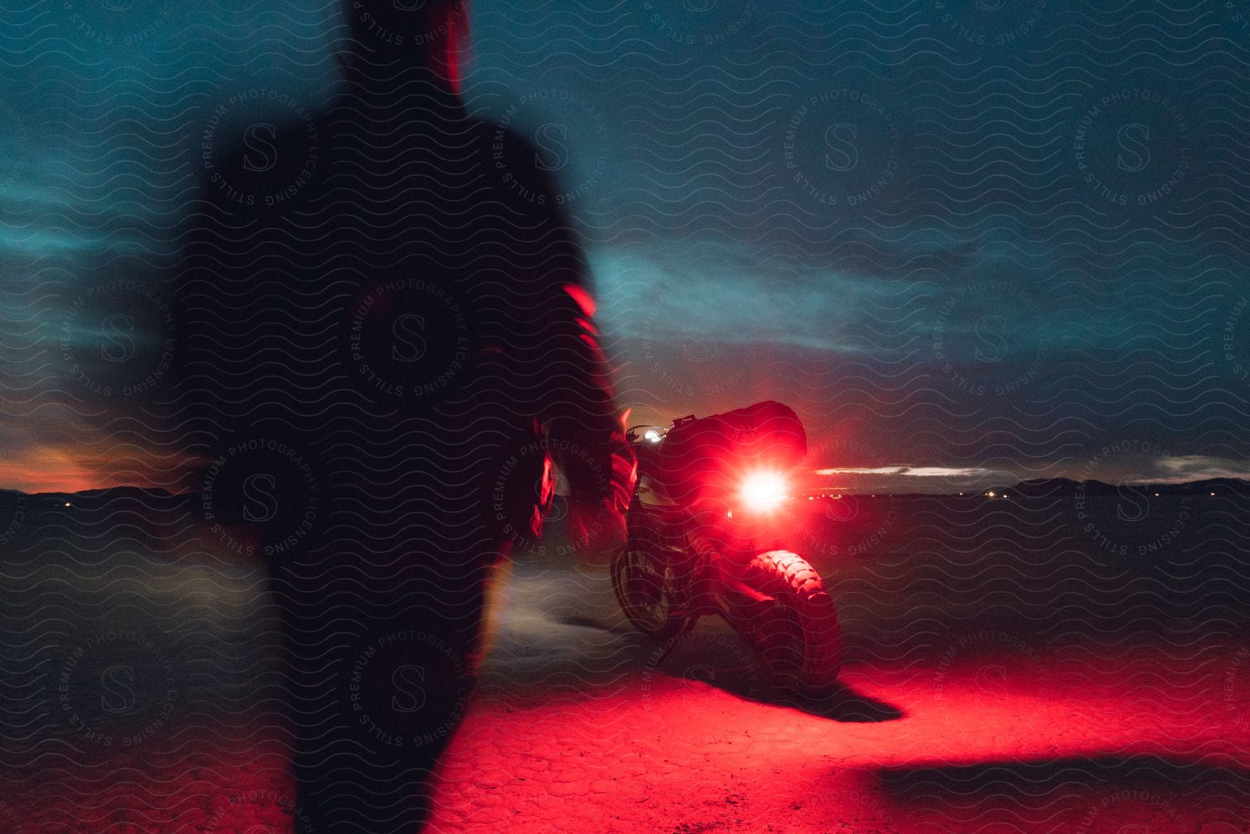 Silhouetted figure with motion blur walking toward a motorcycle with a bright red tail light, set against a dark desert landscape at twilight.