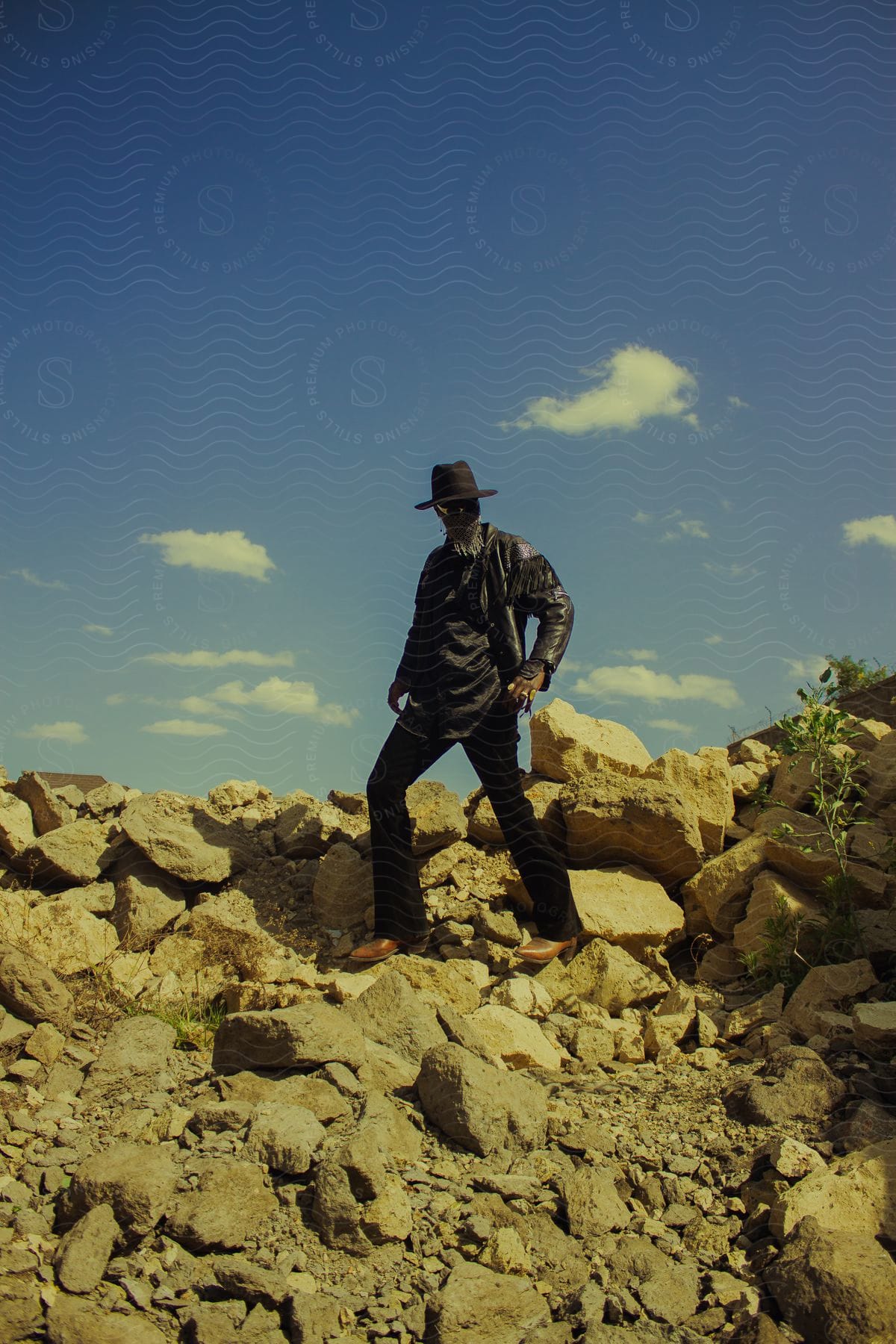Man with brown boots and black clothing including a hat posing on top of rocks in an open-air environment
