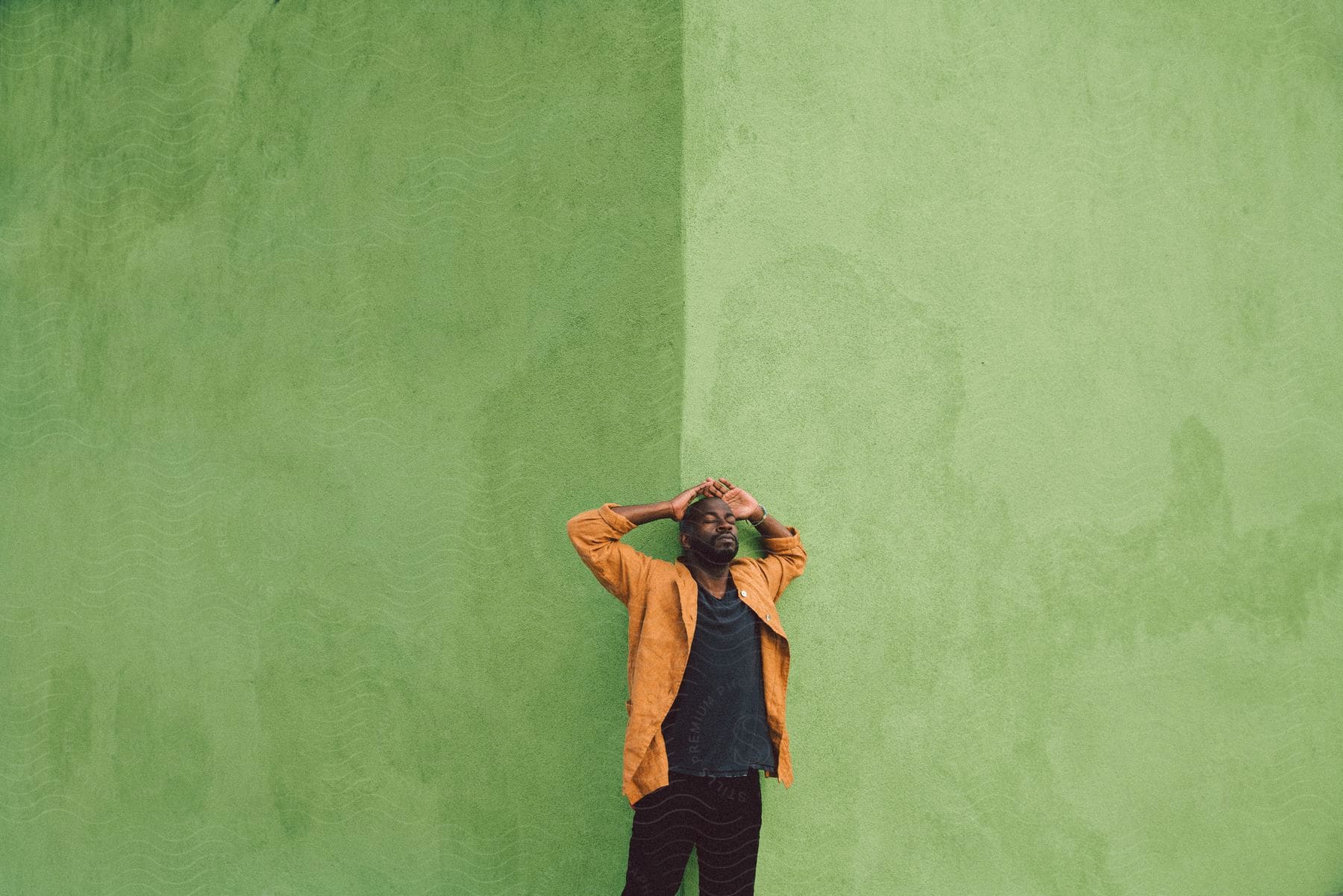 Man standing at the corner of a green building with his hands up over the back of his head