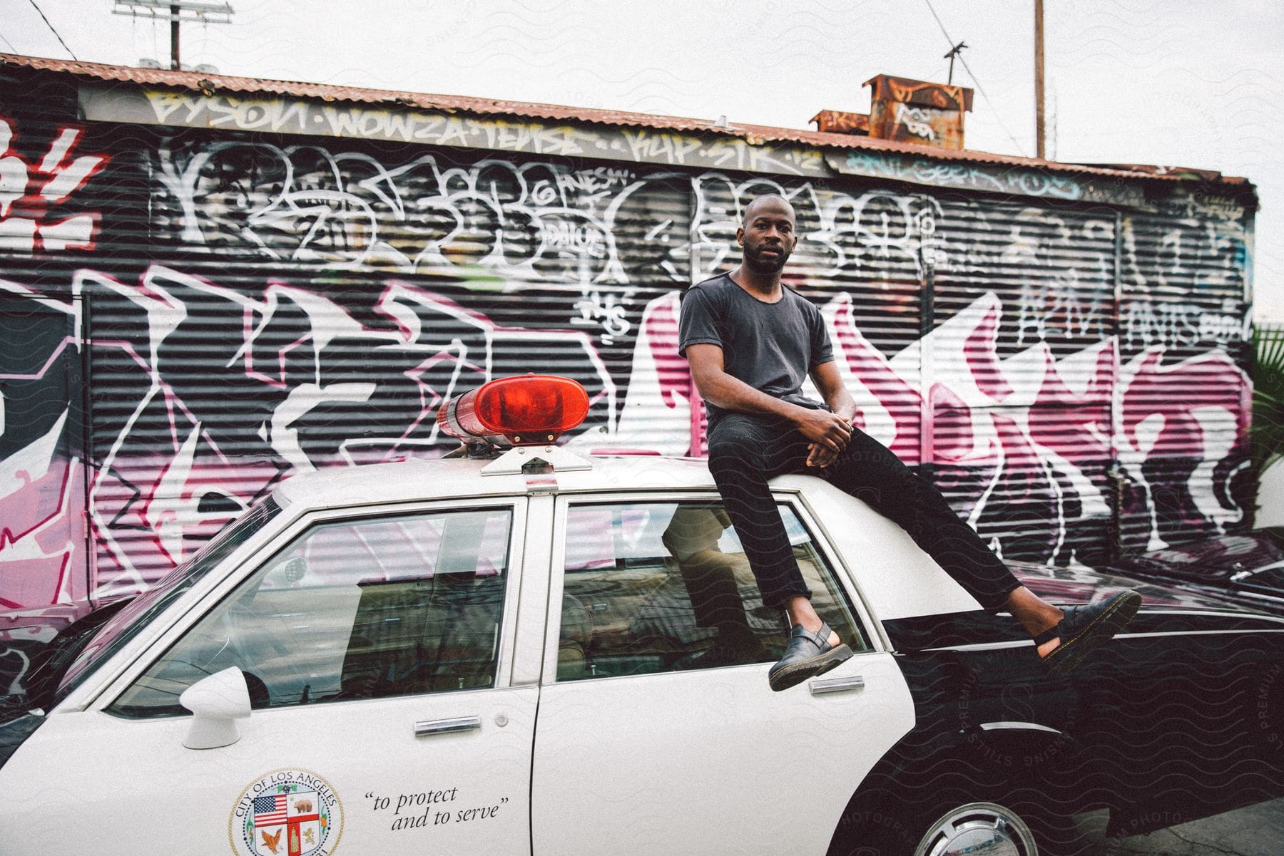 Man sitting on the roof of a police car parked in front of a wall with graffiti.