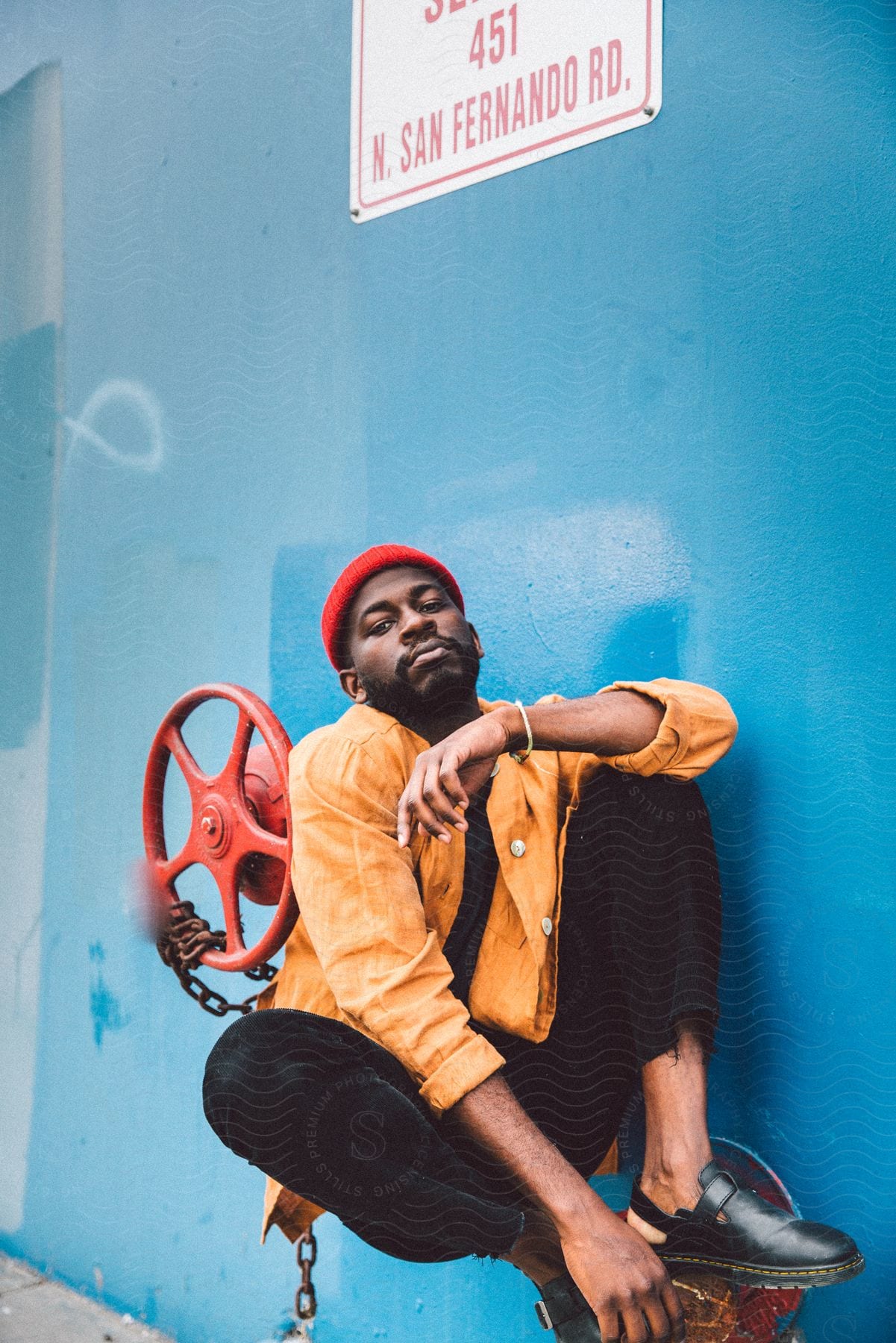 A man in a yellow coat and red cap is sitting on an iron that comes out of a blue wall and there is a valve