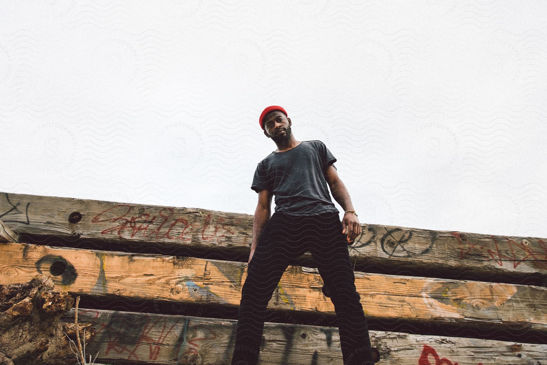 Man with black pants and wearing a red hat posing in an open-air environment with a graffitied wooden wall