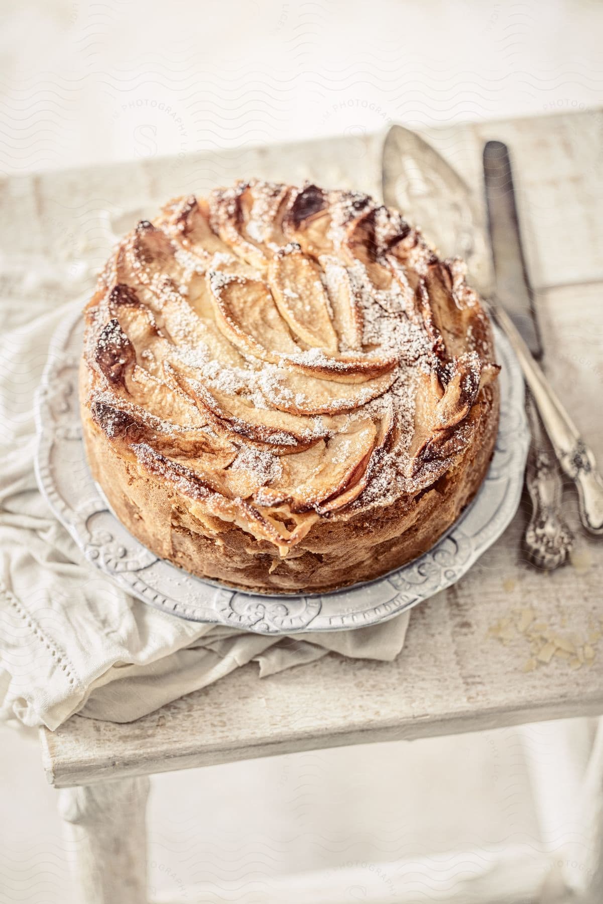 Apple cake dusted with powdered sugar, served on a decorative plate with antique utensils on a rustic white wooden table.