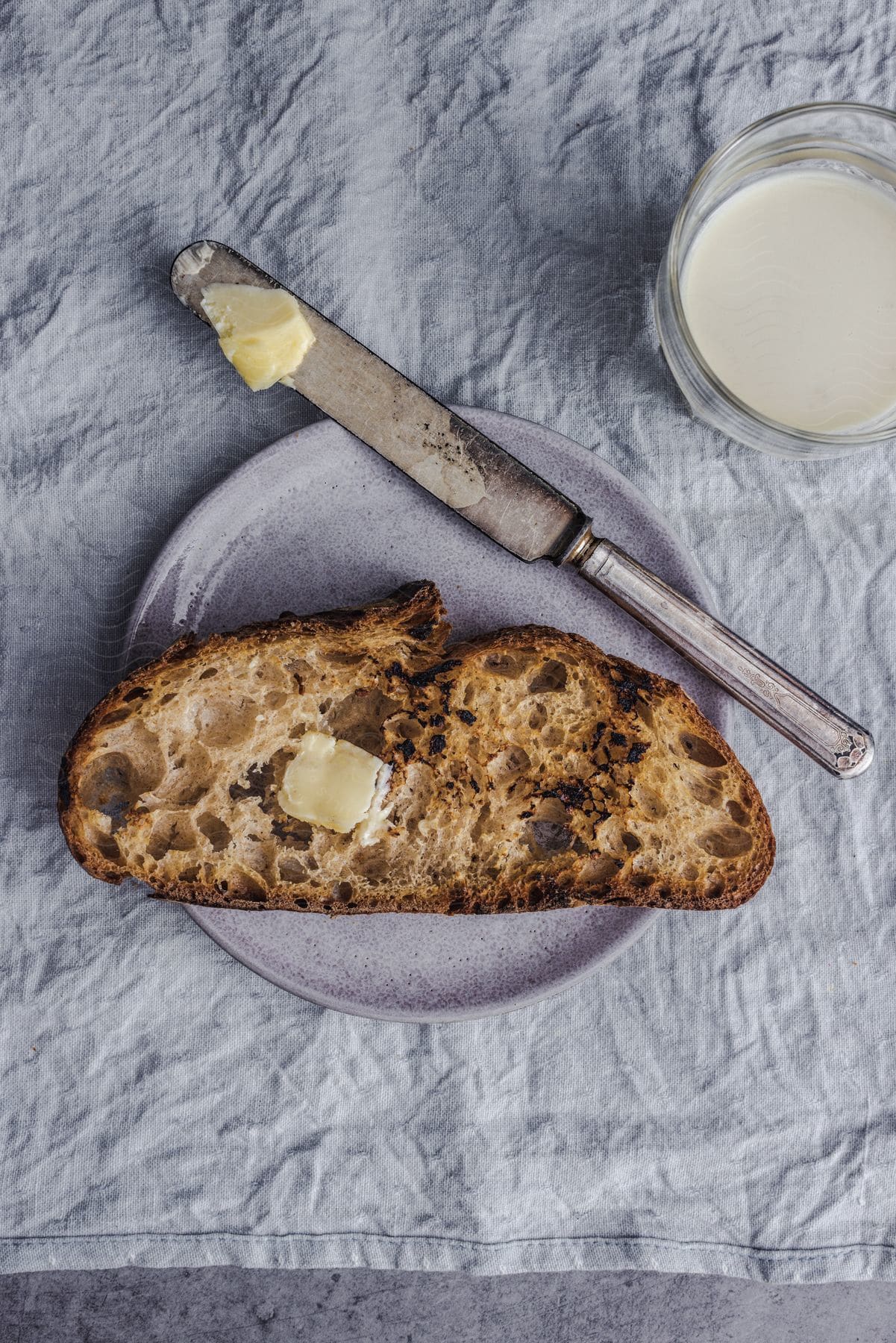 Slice of bread on a plate with butter on a knife and a glass of milk