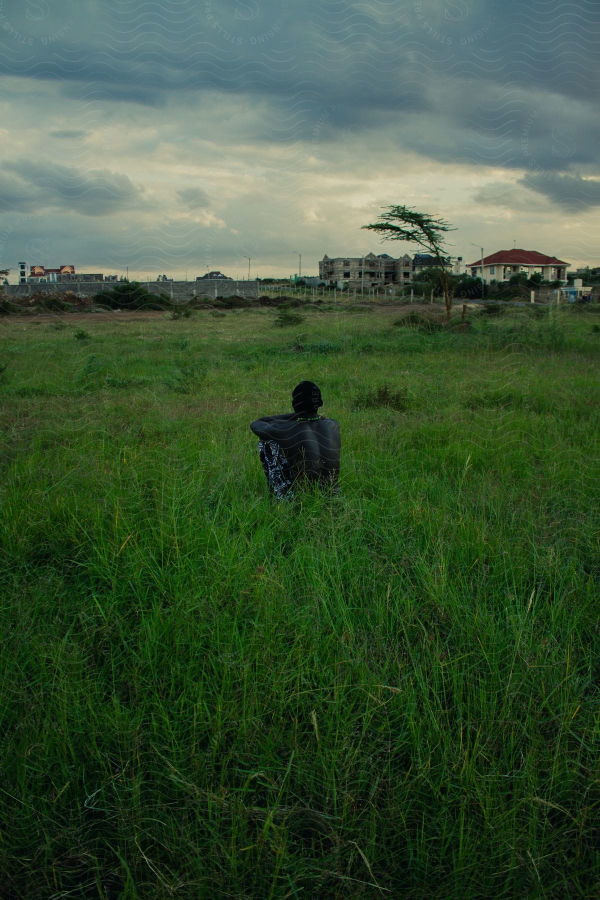 Man with his back without a shirt sitting on a greenish grass overlooking an urban city on a cloudy day