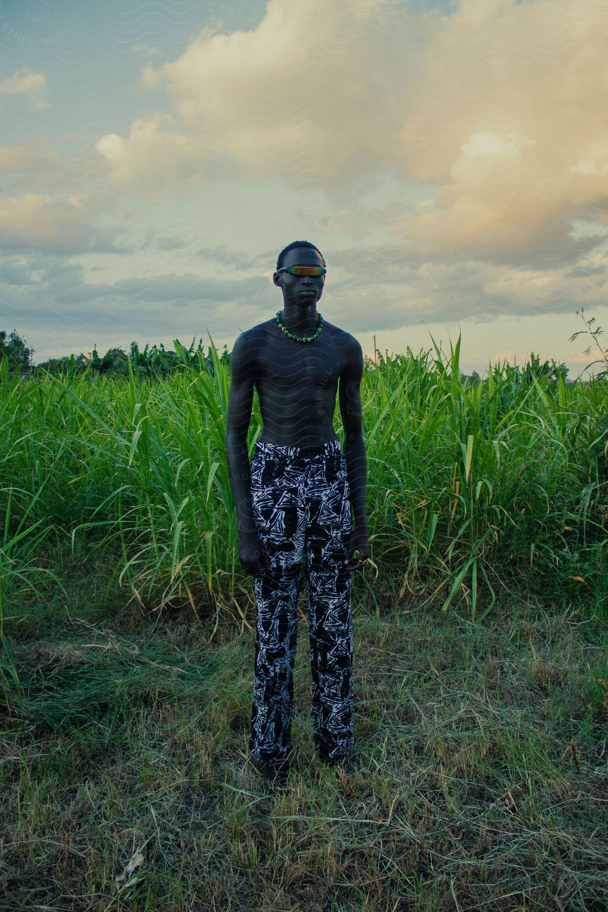 Man without a t-shirt and wearing fashion pants posing with a necklace in futuristic glasses in an agricultural field