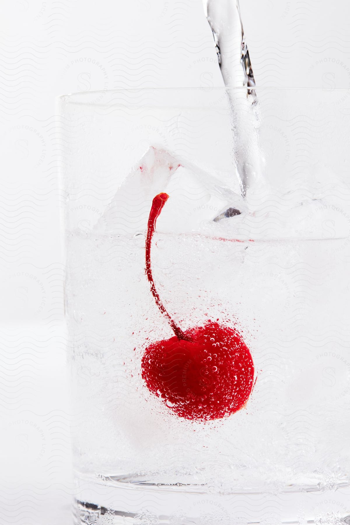 Stock photo of transparent glass with liquid and ice and a submerged cherry with stem.