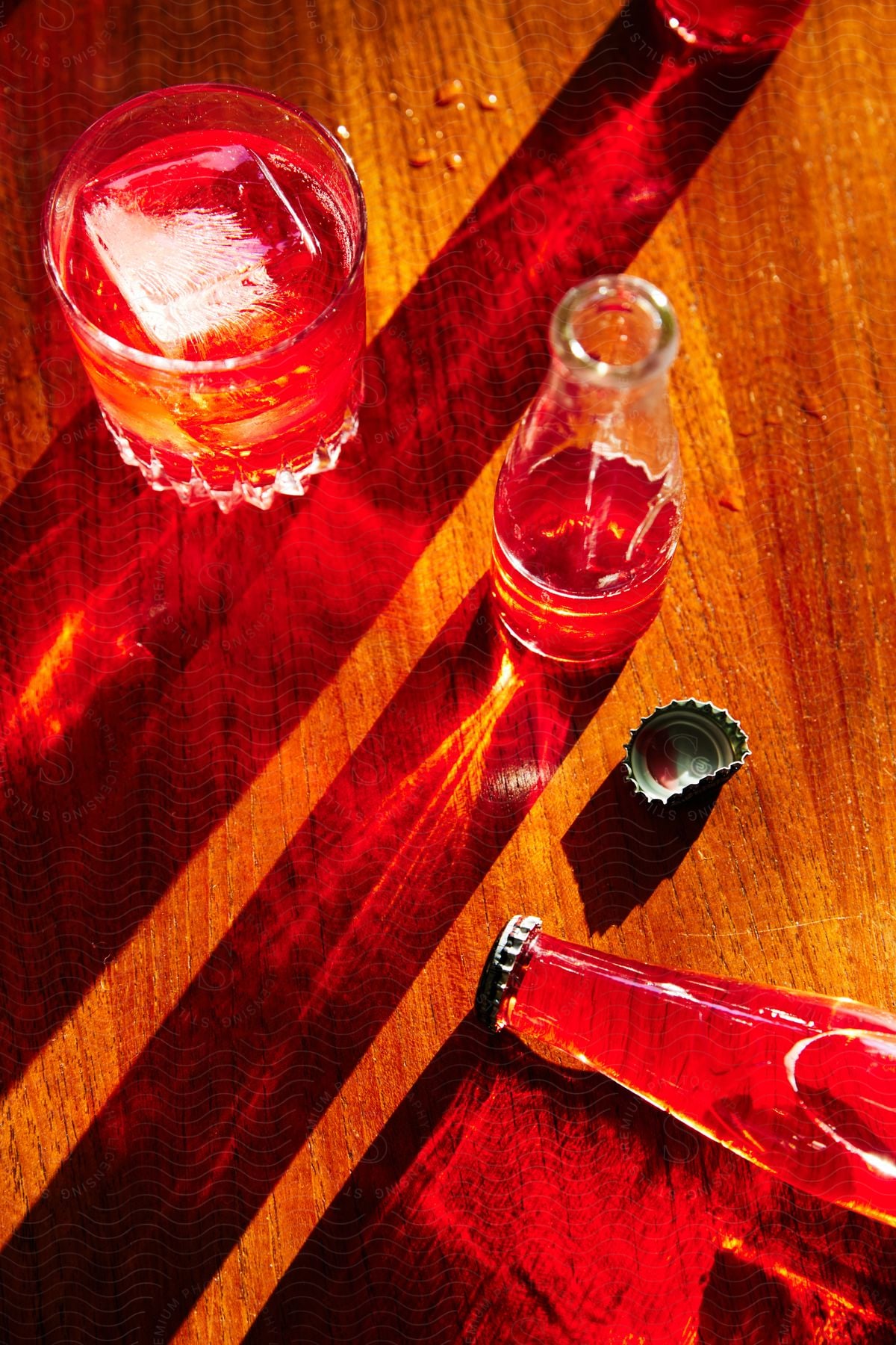 A glass of red liquid with an ice cube and a half-full bottle, near a full bottle of drink, sit on a worn wooden table.