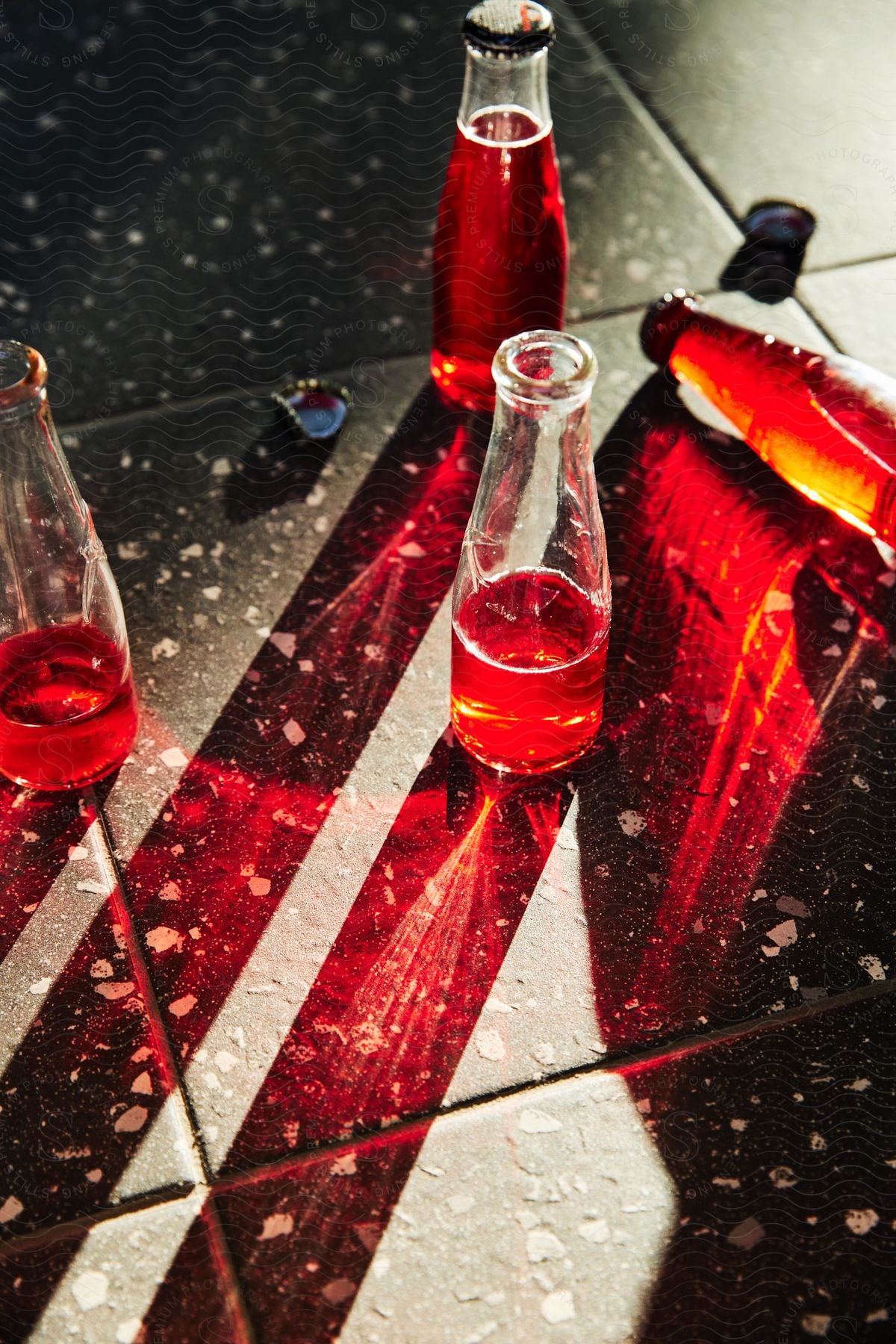 Four glass bottles of red liquid on a speckled surface, with sunlight casting vivid red reflections and shadows.