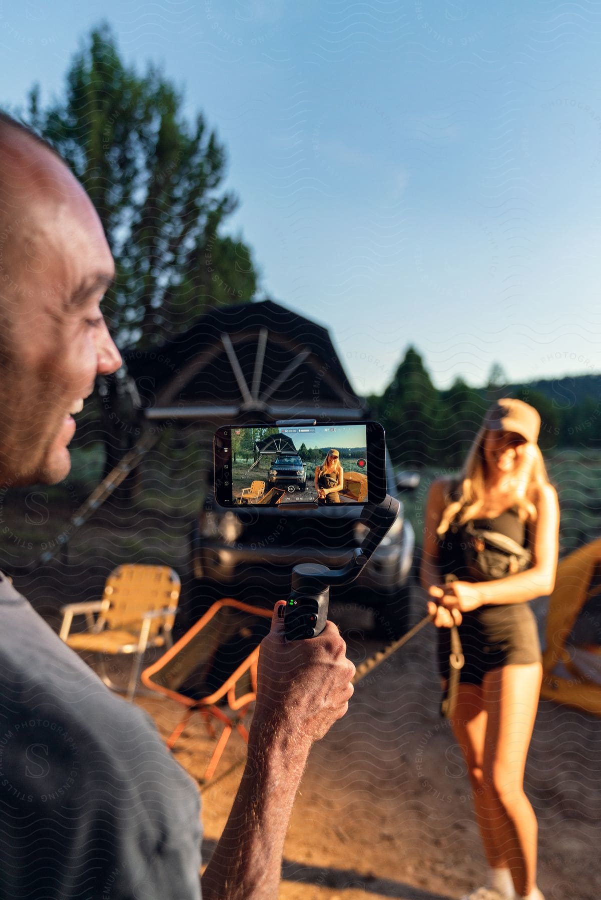 a man filming a woman wearing a cap, a black top, and shorts with a smartphone near a tent in the field.