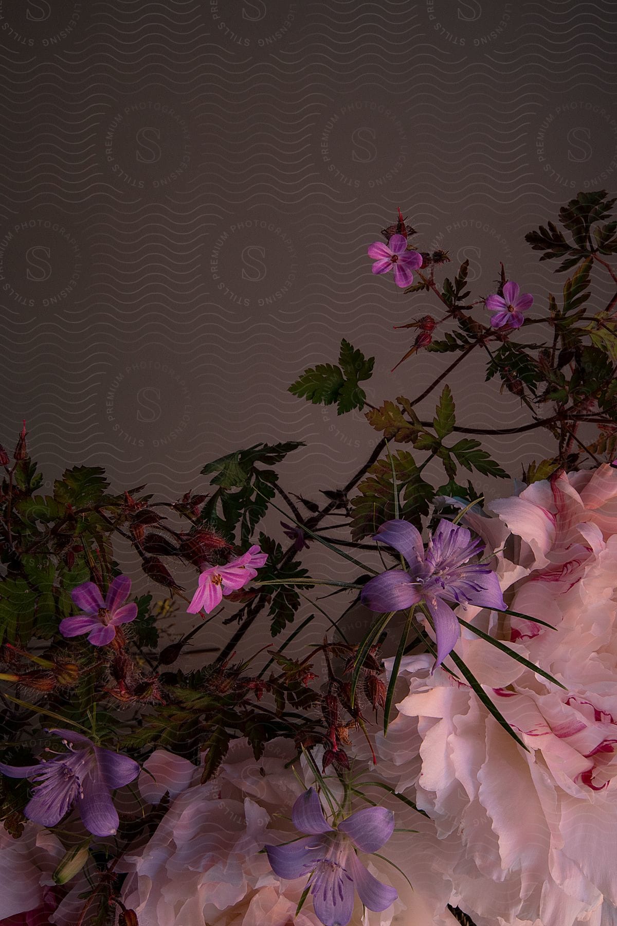 Against a muted grey backdrop, pink lilies, delicate violet blooms, and white and pink carnations create a harmonious floral arrangement.