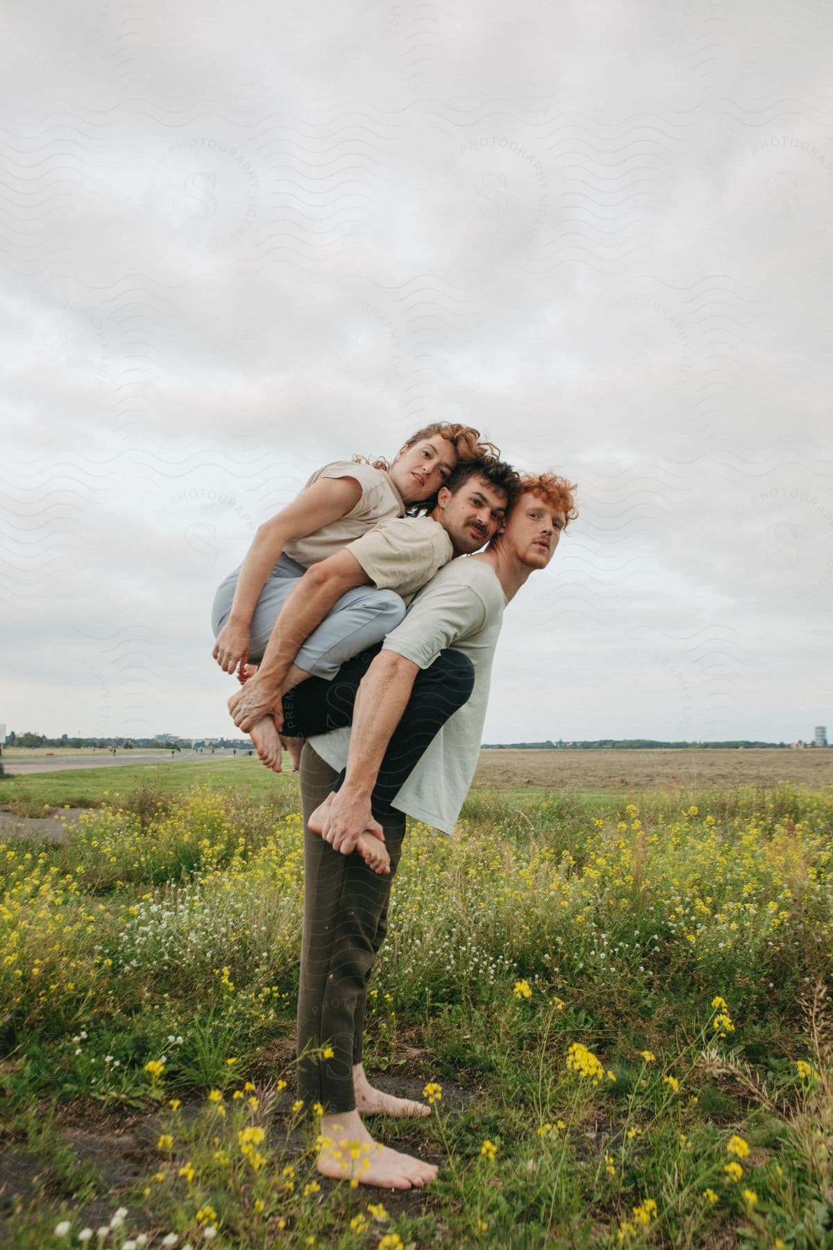 Three people outdoors, one standing barefoot in a field of yellow flowers, while the other two are perched on his back.