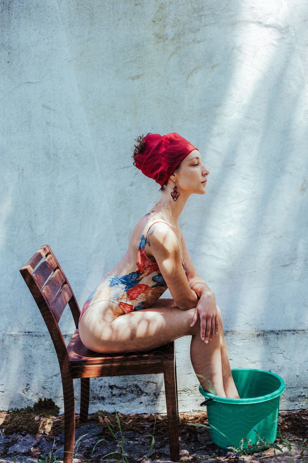 Stock photo of a woman wearing a red hat, earrings, and a colorful swimsuit sitting on a wooden chair with her feet in a turquoise bucket.