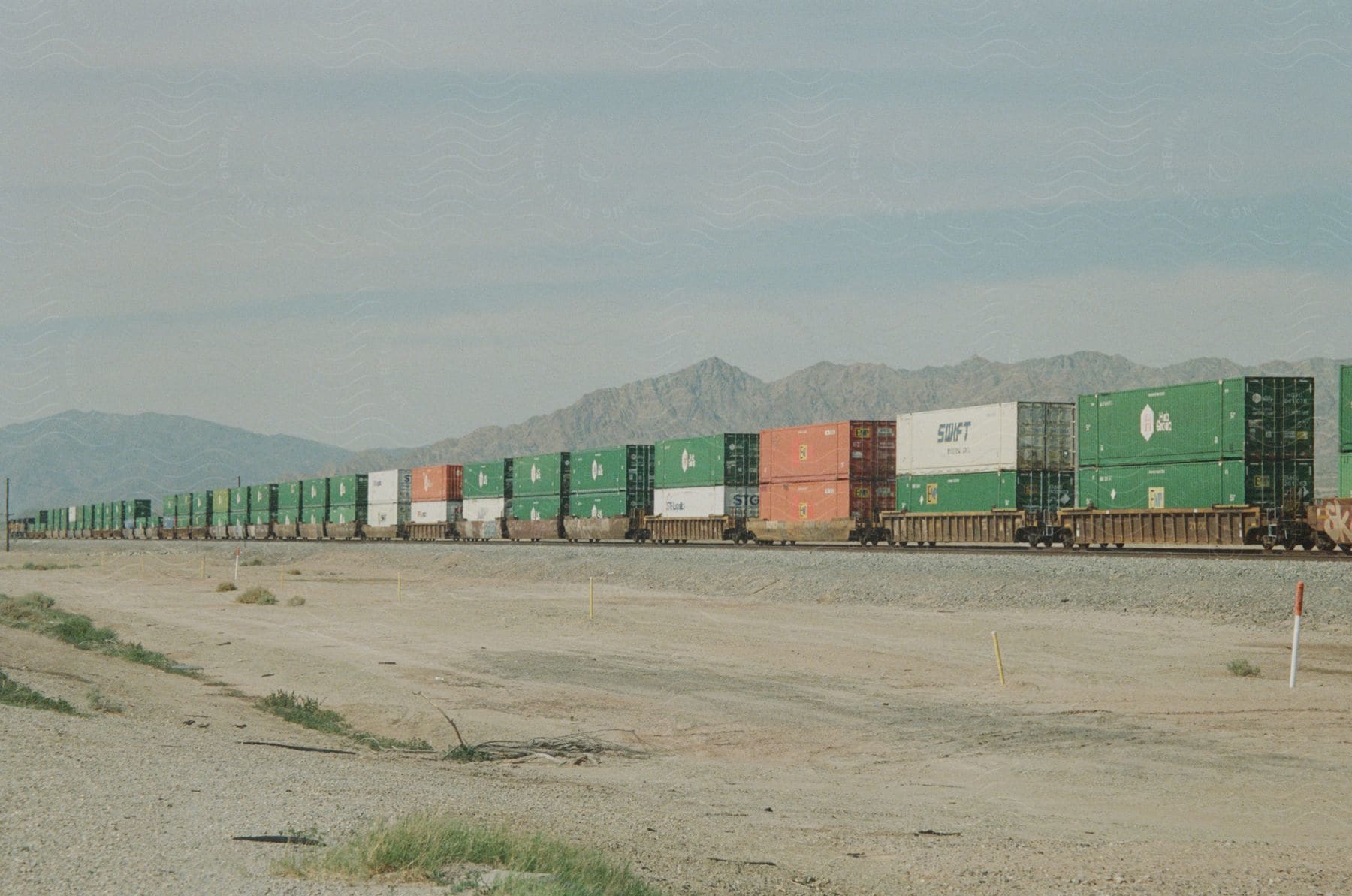 Long freight train with several colorful containers in a desert area with mountains in the background.