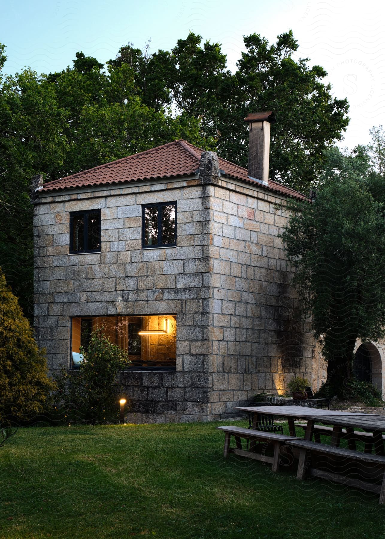 A house made of stone and in front of it is a yard with wooden outdoor tables and behind the house there are some trees