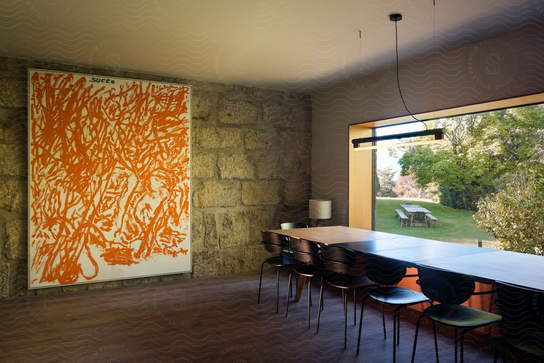 Painting on the wall and a long table and chairs facing a large window with a view of a picnic table and trees outside