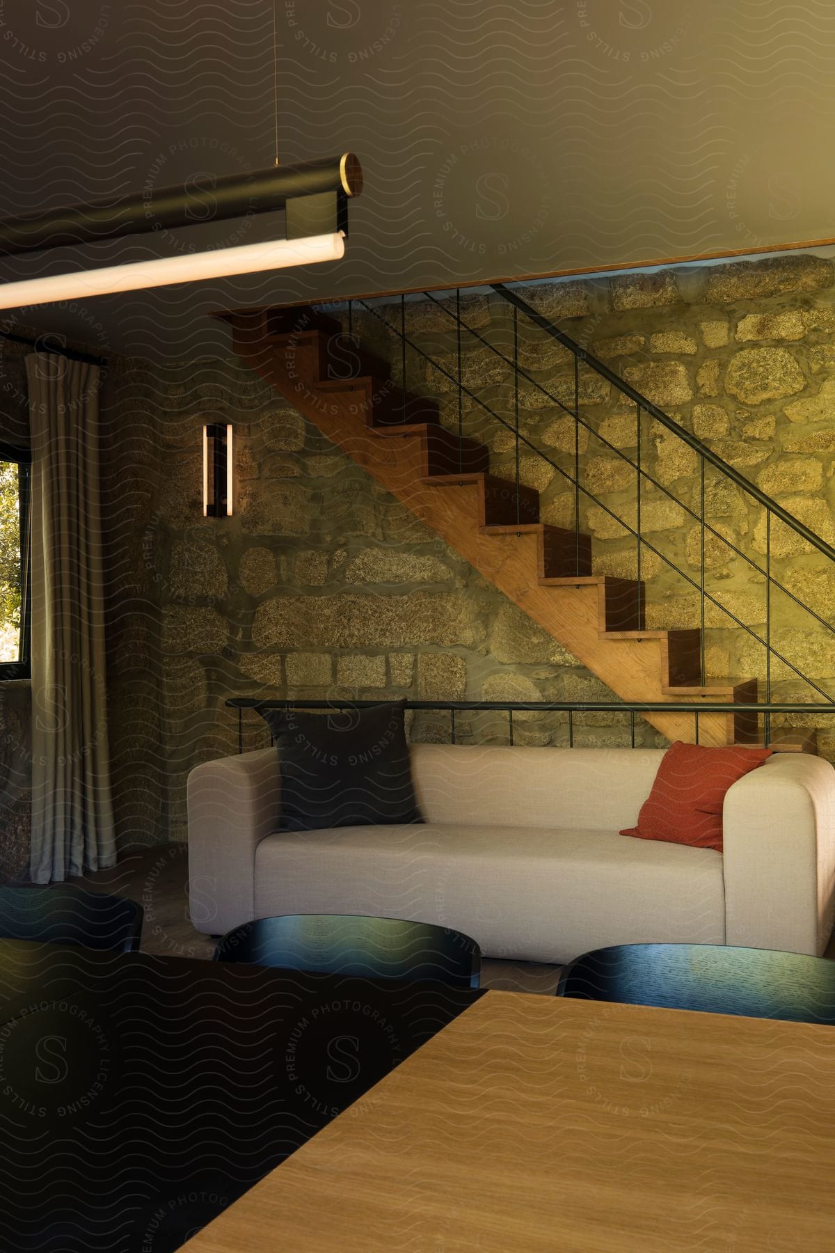 Living room with a beige sofa adorned with black and orange cushions. Above the sofa, a wooden staircase with a minimalist black railing ascends against a rustic stone wall.
