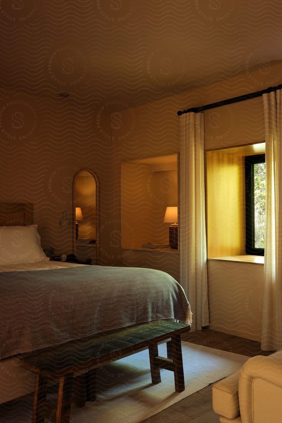 a wooden platform bed with white bedding and a woven bench at its foot. Sunlight streams through a large window behind the bed