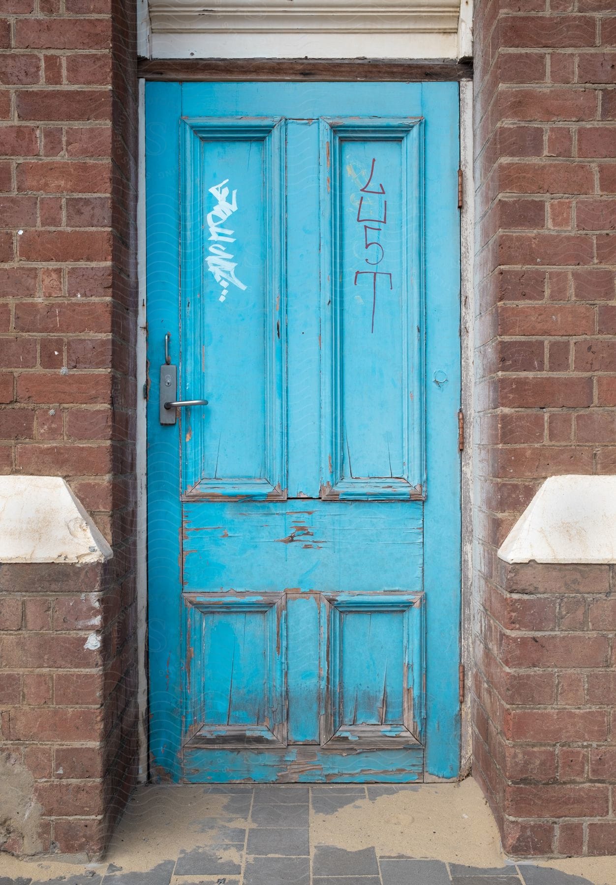 An old blue door with deterioration and some writing marks and it is on a brick wall