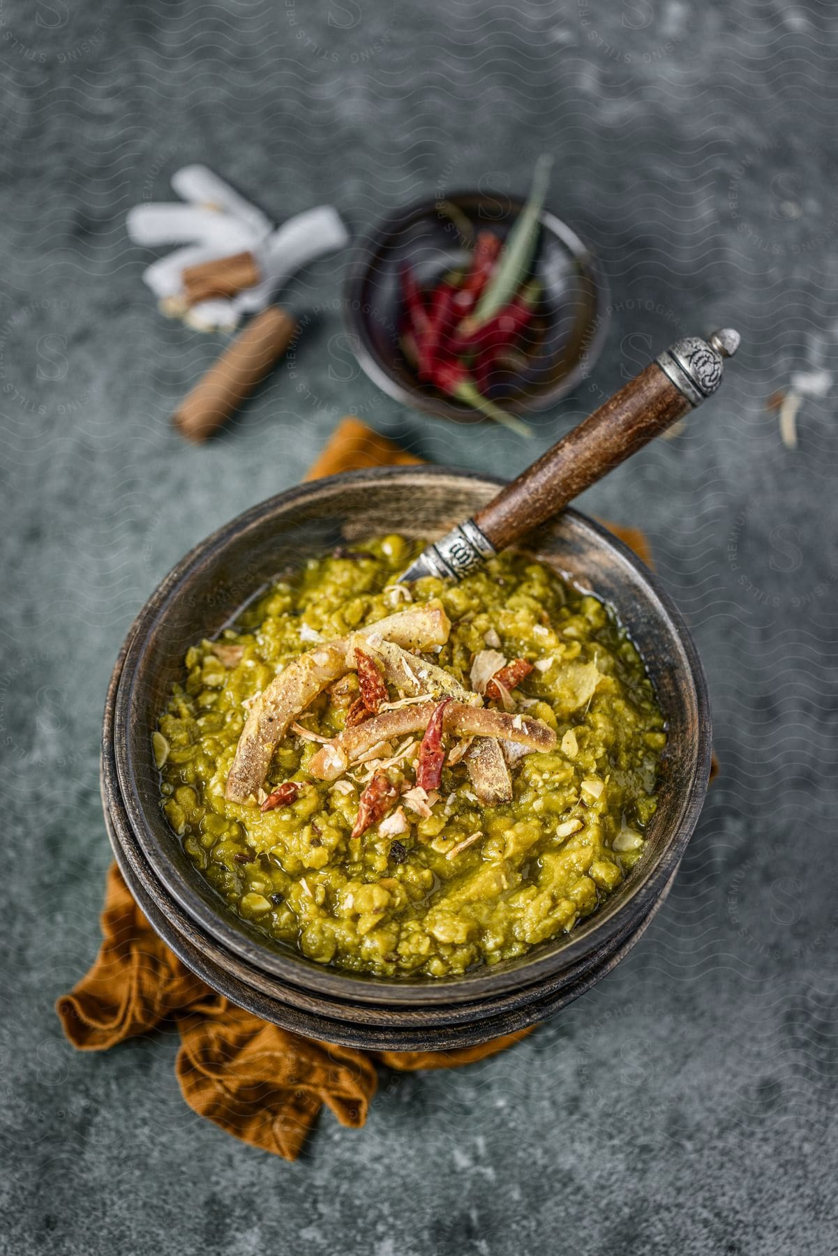 A bowl filled with a thick green curry topped with crispy fried shallots and red chilies. The bowl sits on a textured gray surface with a brown cloth underneath.