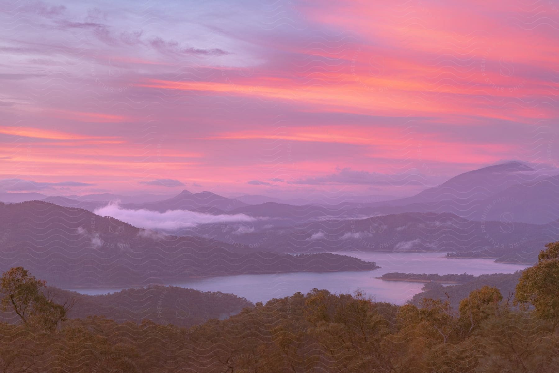 A scenic landscape at dawn or dusk, with vibrant shades of pink and purple in the sky, mist over wooded mountains and a calm lake reflecting the colors of the sky.