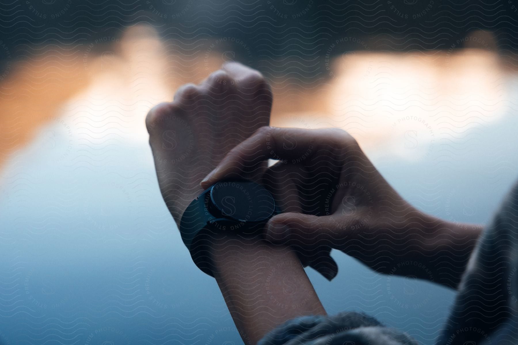 A woman is looking down at her wrist, where a black smartwatch is, near the lake at dusk.