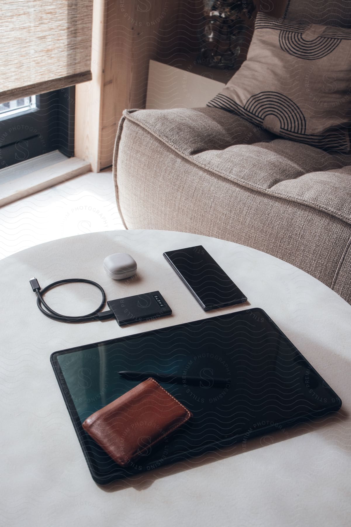 a wooden coffee table with a black tablet computer, a black phone, a brown leather wallet, a black pen, and a white charging case for earbuds on it.