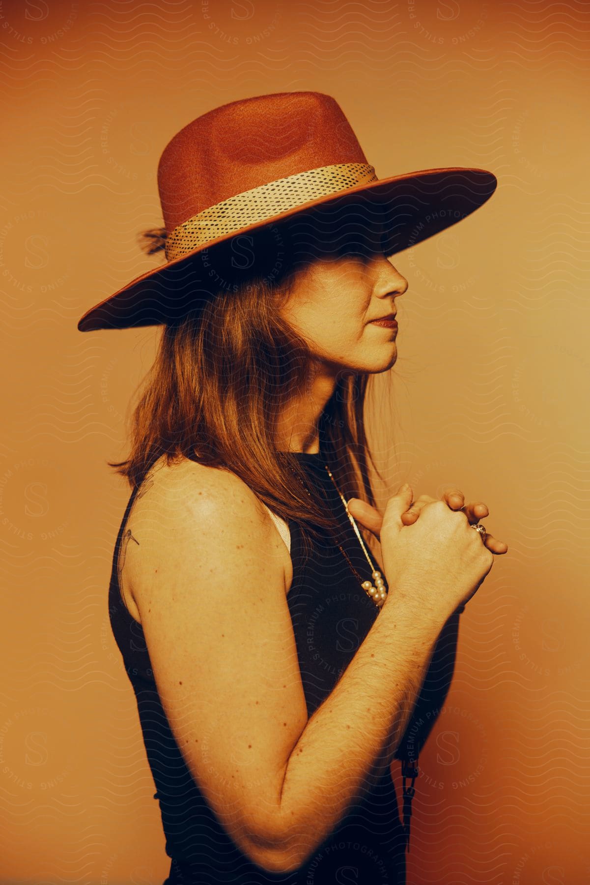 A woman with long brown hair wearing a sun hat stands with her hands clasped in front of her and turned to the side