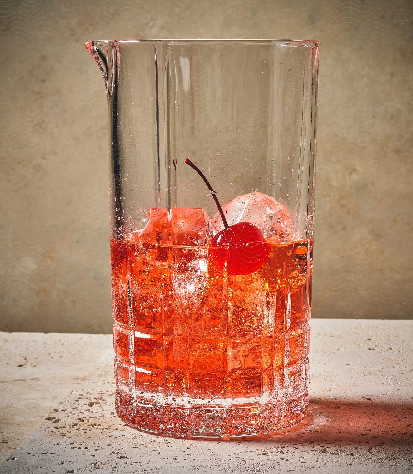 A red alcoholic drink with ice and a cherry in a tall glass on a marble table