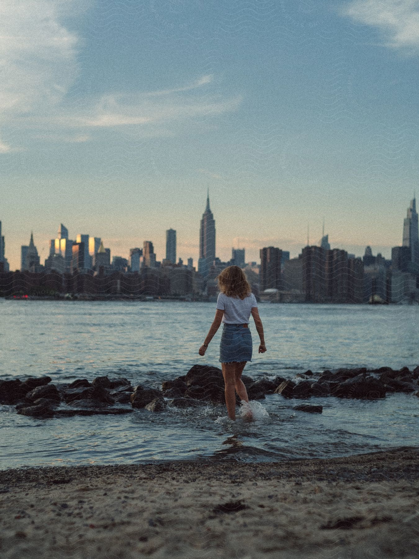 A person walking along the shoreline of a city beach with skyscrapers in the background under a sunny sky