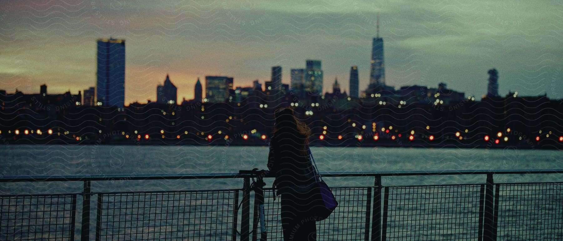 Woman in black overcoat leans against handrails watching coastal city with skyscrapers