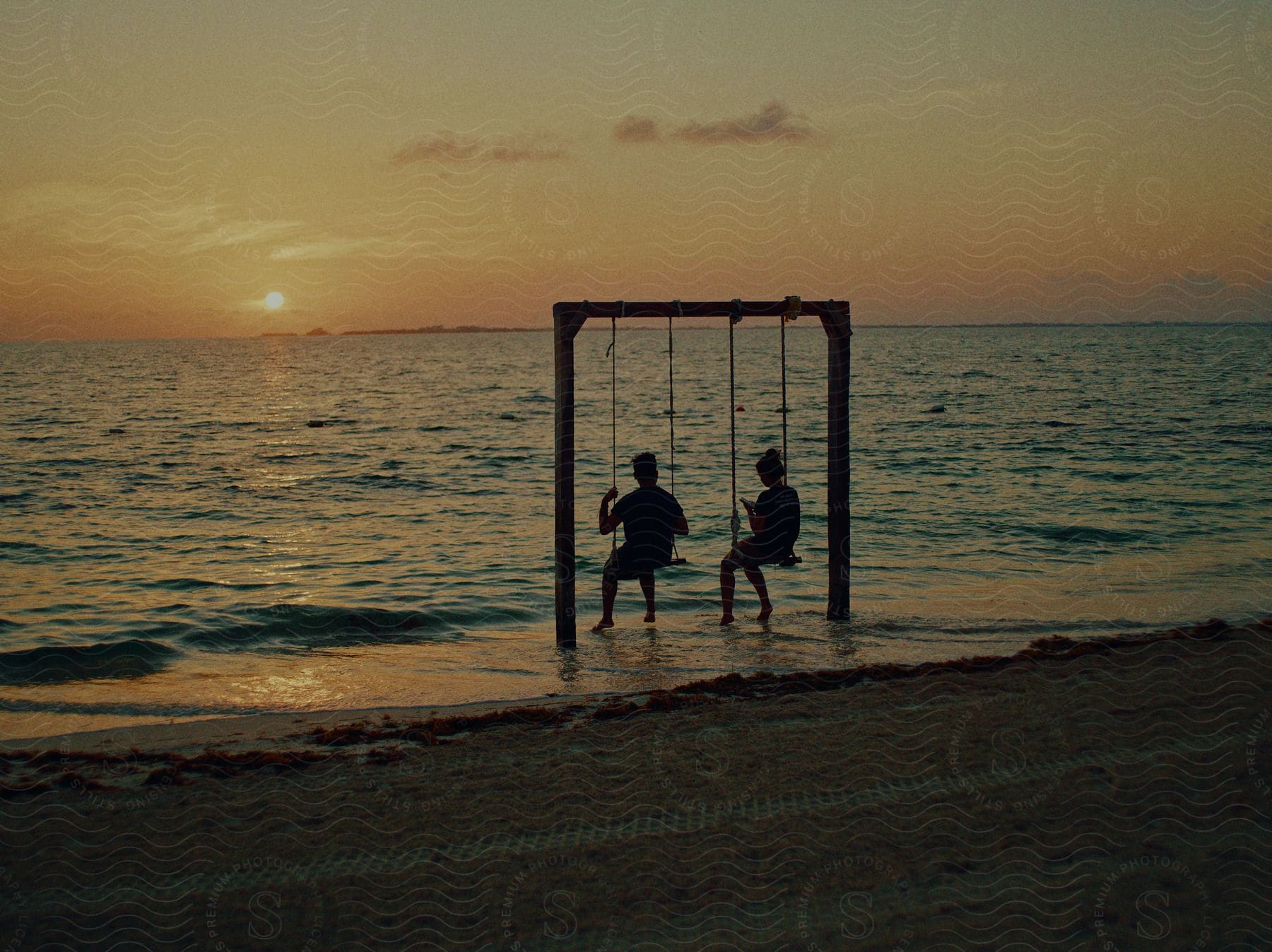 Two boys sitting with their backs to a seashore swing against the light on a beach at dusk