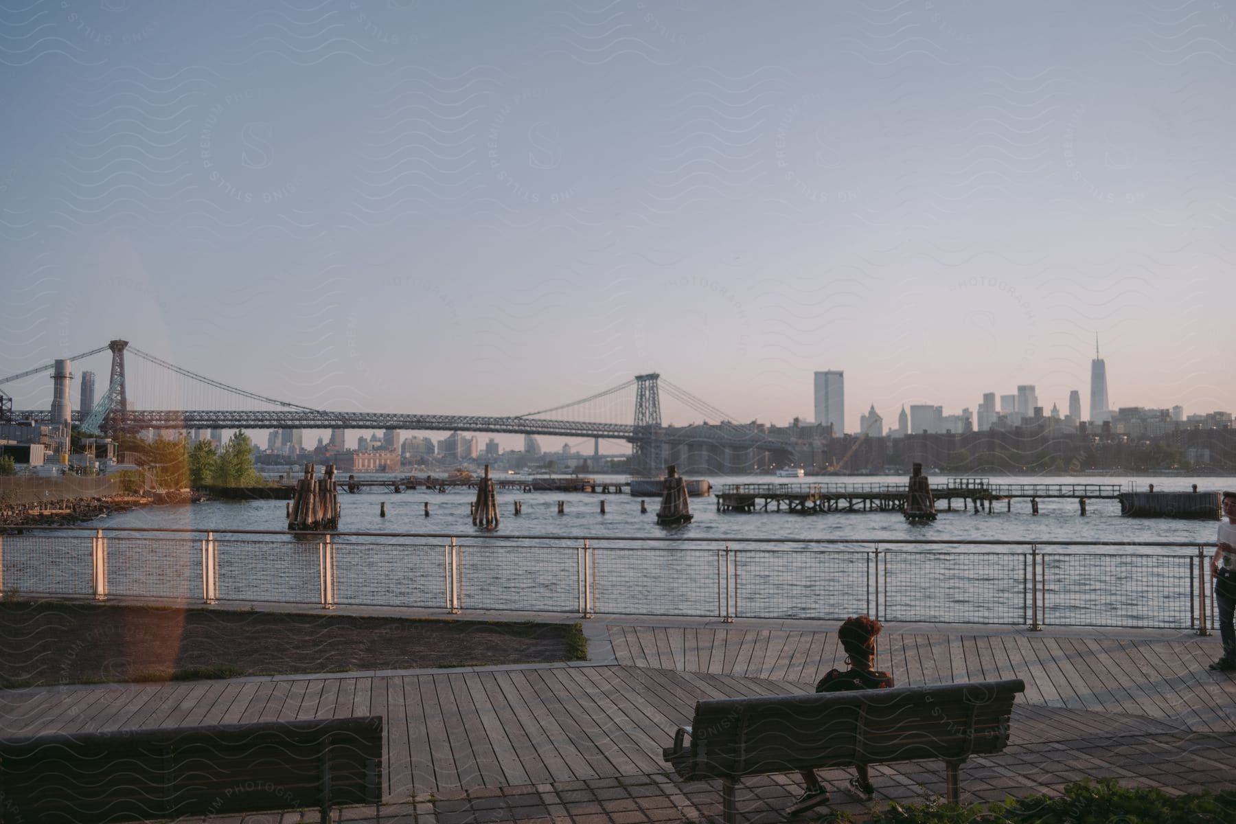 A woman on a bench at the pier looking out at boats and a long bridge connecting to the city in the horizon at dawn